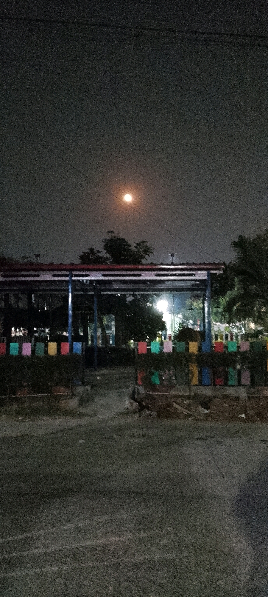 The image shows a nighttime scene in Satriamekar, Indonesia, featuring a building with a red roof supported by blue pillars, possibly a pavilion or a covered walkway. The building is fronted by a low hedge decorated with colorful square panels in red, yellow, blue, and green. Beyond the hedge, a paved area suggests a walkway or road. In the background, trees are visible, and a bright, full moon illuminates the dark sky, casting a soft glow over the scene. The overall atmosphere is quiet and still, characteristic of a late hour.