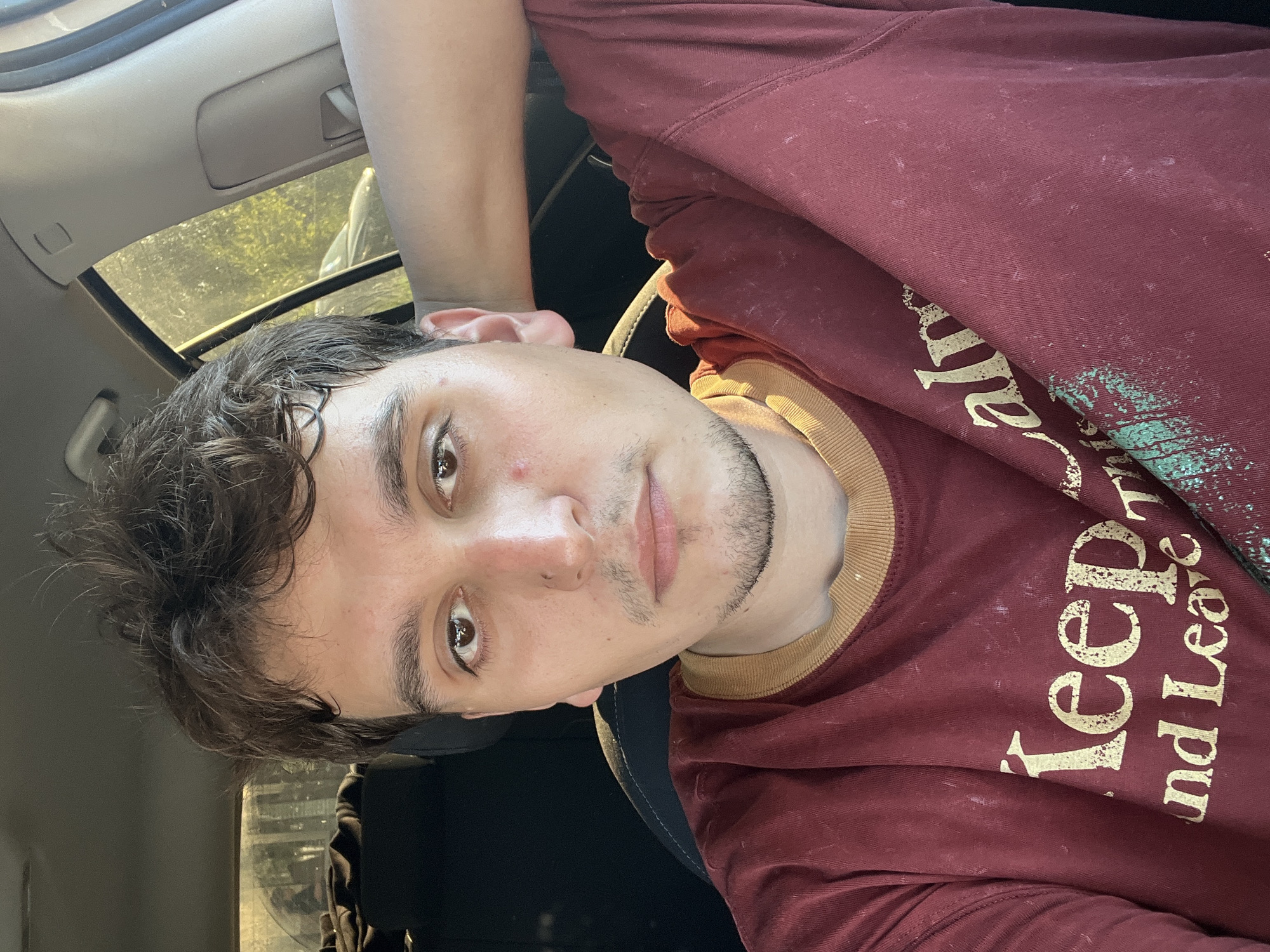 A medium shot shows a young man with dark, messy hair sitting in the driver's seat of a car, looking at the camera. He is wearing a maroon t-shirt with a yellow collar and a faded gold design that reads "Keep Calm and Leave." His left arm is resting behind his head. The background shows the interior of the car and a blurred view of greenery and a car through the window, suggesting an outdoor location. The image is a selfie taken from a slightly low angle.