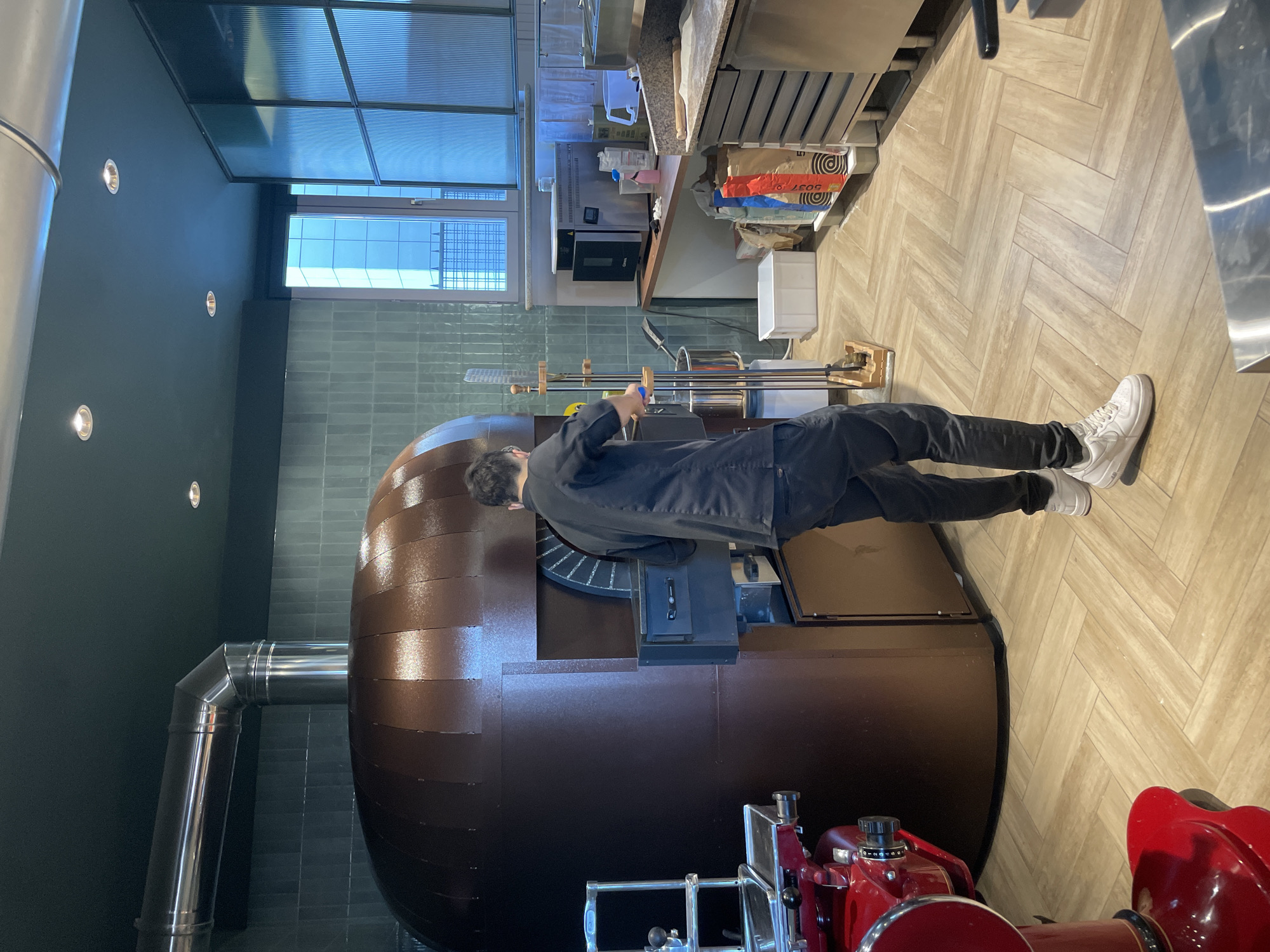 The image shows an interior setting, presumably a restaurant or kitchen, in Busto Arsizio, Italy. A man dressed in black stands in front of a large, rounded, brown pizza oven. He is holding long-handled tools, likely used for manipulating food inside the oven. To the right of the oven, a red meat slicer is visible. The floor is tiled in a herringbone pattern. Above the oven, there is a work surface with kitchen equipment and supplies. A window lets in natural light. A stainless steel chimney runs up the side of the oven. The walls are dark green with recessed lighting.