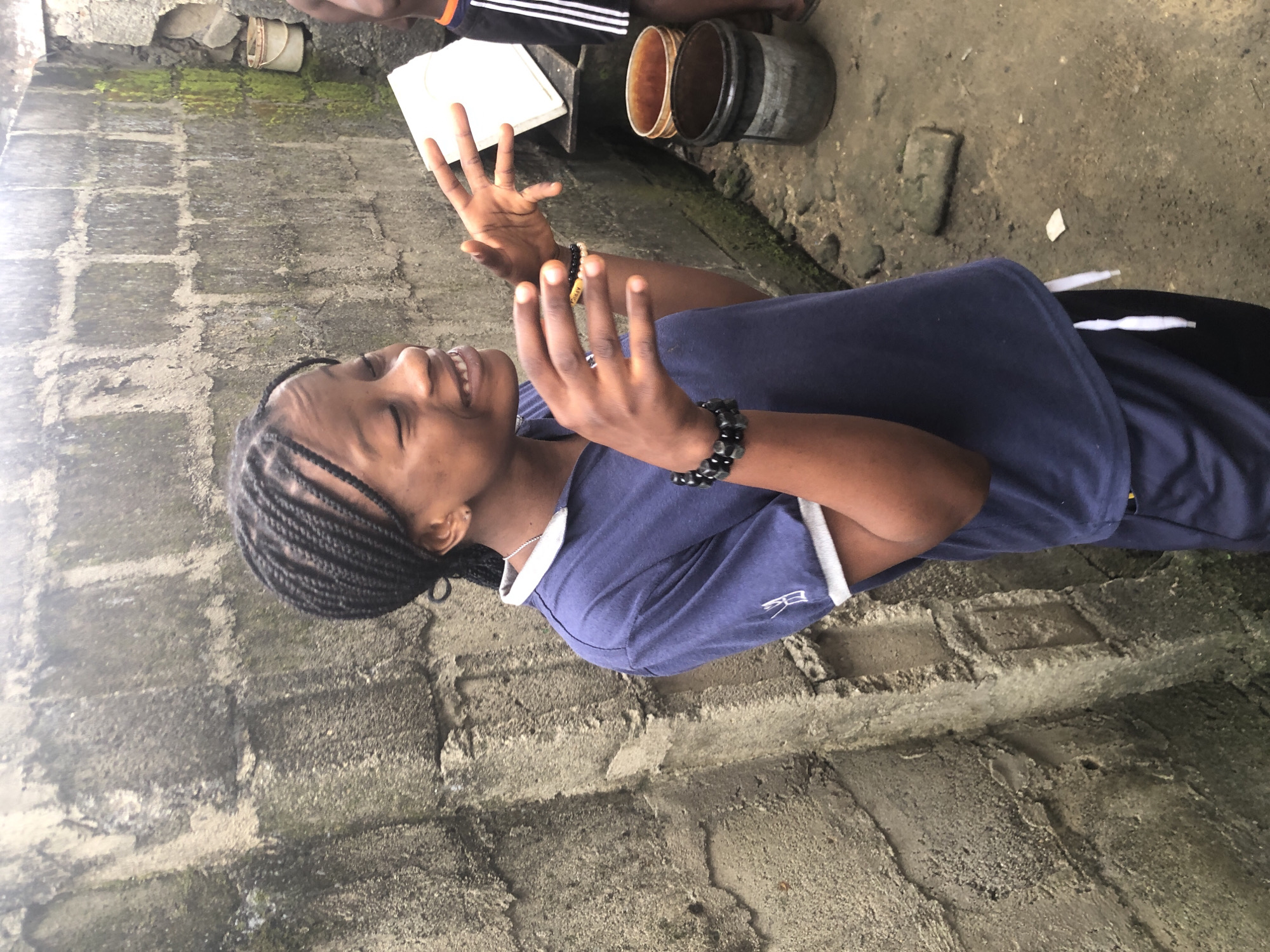 A young girl with braided hair is pictured in what appears to be an outdoor urban environment in Ado, Nigeria. She is wearing a blue polo shirt with white trim at the collar and sleeves, and dark athletic shorts with white drawstring. On her left wrist, she wears a black beaded bracelet. Her expression is animated, with eyes closed and mouth open, as if she is speaking or laughing, and her hands are raised in a gesture of explanation. Behind her, a rough, grey concrete wall with visible brickwork and patches of green moss forms the backdrop. To her right, a white object, possibly a book or board, rests on a surface, and further back, two buckets are visible, one orange and one black. The ground is dirt and appears damp in places. The overall scene suggests a casual, possibly residential setting.