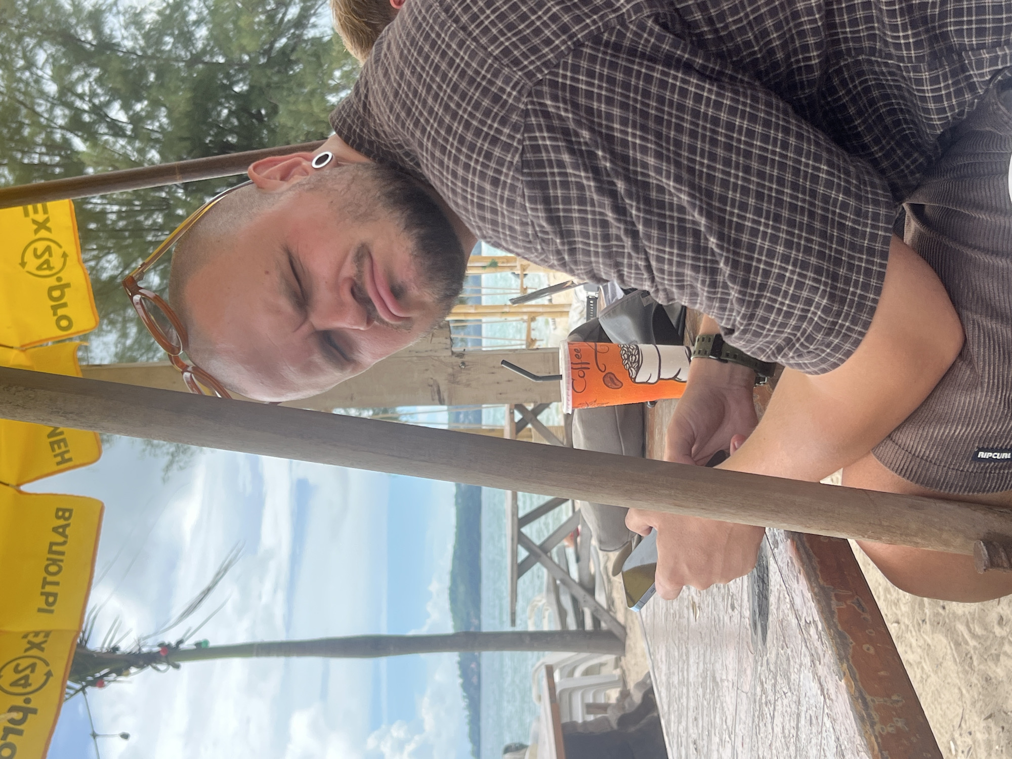 A man is seated at a wooden table on a beach, looking towards the camera with a grimace. He has a shaved head, a beard, and is wearing sunglasses on his head and an earring in his left ear. His right arm is resting on the table, holding a smartphone. A cup of coffee with a straw sits on the table next to him. Behind him, the ocean and a distant island are visible. The scene appears to be in Choeng Thale, Thailand.
