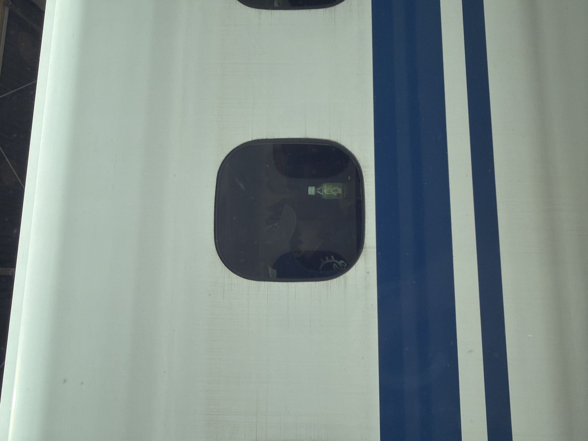 The image shows a close-up of the exterior of a white train car with blue stripes. There are two rounded, dark windows visible. Inside the left window, a small green bottle of green tea is clearly visible, along with a reflection that suggests a person is sitting inside. The train appears to be stationary, possibly at a station in Nagoya, Japan. The environment is clean and well-maintained, consistent with Japanese public transportation.