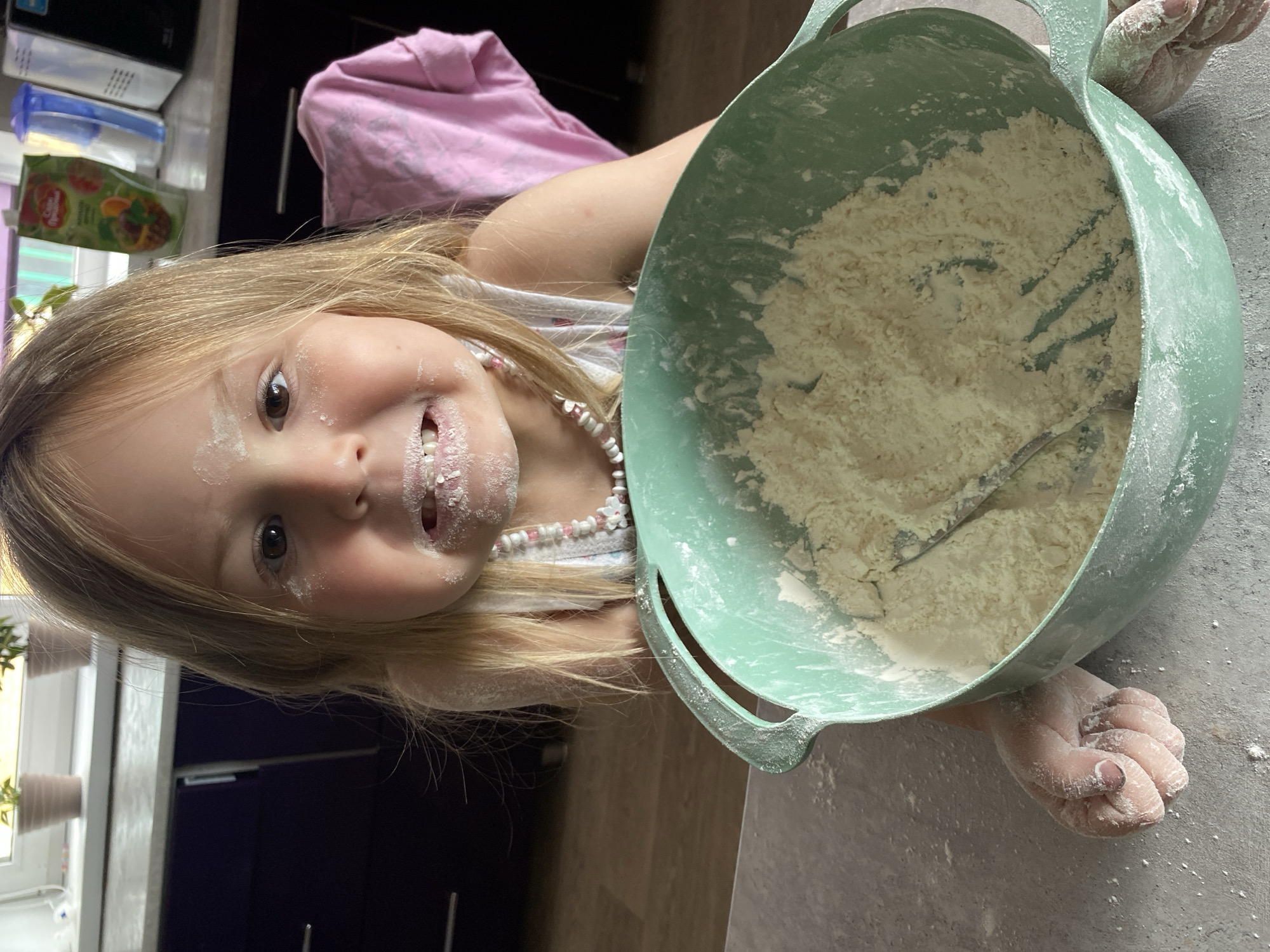 A young girl with blonde hair, covered in flour, makes a funny face as she looks into the camera. She is in a kitchen, with a bowl of flour and a whisk in front of her. A pink shirt is visible in the background, along with kitchen cabinets and a window. The location is in the city of Chaikovsky, Russia.