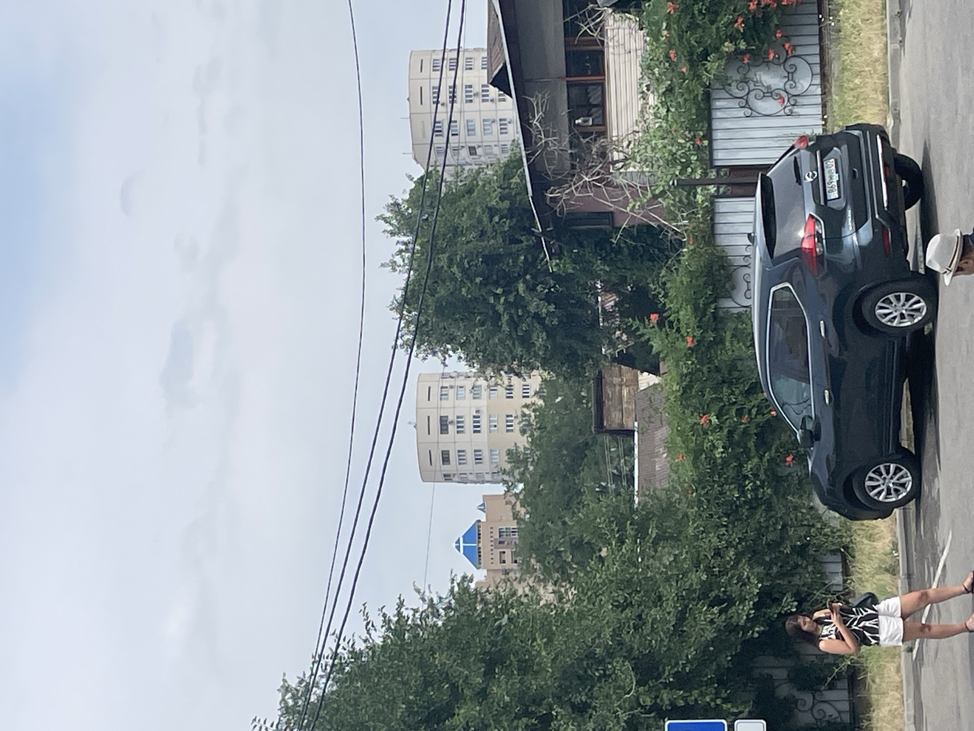 An overcast day in Almaty, Kazakhstan, with several trees obscuring the view of apartment buildings in the background. A dark-colored SUV is parked in the foreground, partially obscuring a woman standing on the left, who is looking at her phone. The woman is wearing a black and white patterned sleeveless top and white shorts. A man wearing a white hat is visible in the lower right corner of the image. The scene appears to be a residential area with a street, a sidewalk, and a fence with decorative elements.