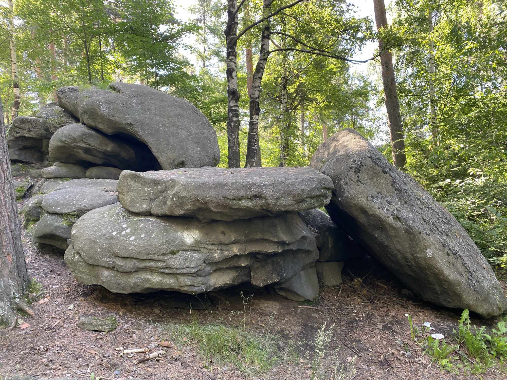 The image depicts a cluster of large, weathered boulders in a forest setting. The boulders are grey and appear to be composed of granite or a similar rock type, with some showing signs of moss and lichen growth. They are arranged in a haphazard manner, with some stacked precariously on top of others, creating nooks and crannies. One particularly large, elongated boulder is leaning against the cluster. The ground around the boulders is covered with a mixture of dry leaves, pine needles, and small twigs. There is sparse green grass growing in patches, particularly near the base of the rocks. Behind the boulders, a forest of deciduous and coniferous trees is visible, with sunlight filtering through the canopy, dappling the scene with light and shadow. The overall impression is one of a natural, undisturbed environment. The location is specified as the Ural Mountains near Yekaterinburg, Russia, a region known for its granite rock formations and forests. There are no people or animals visible in the image.