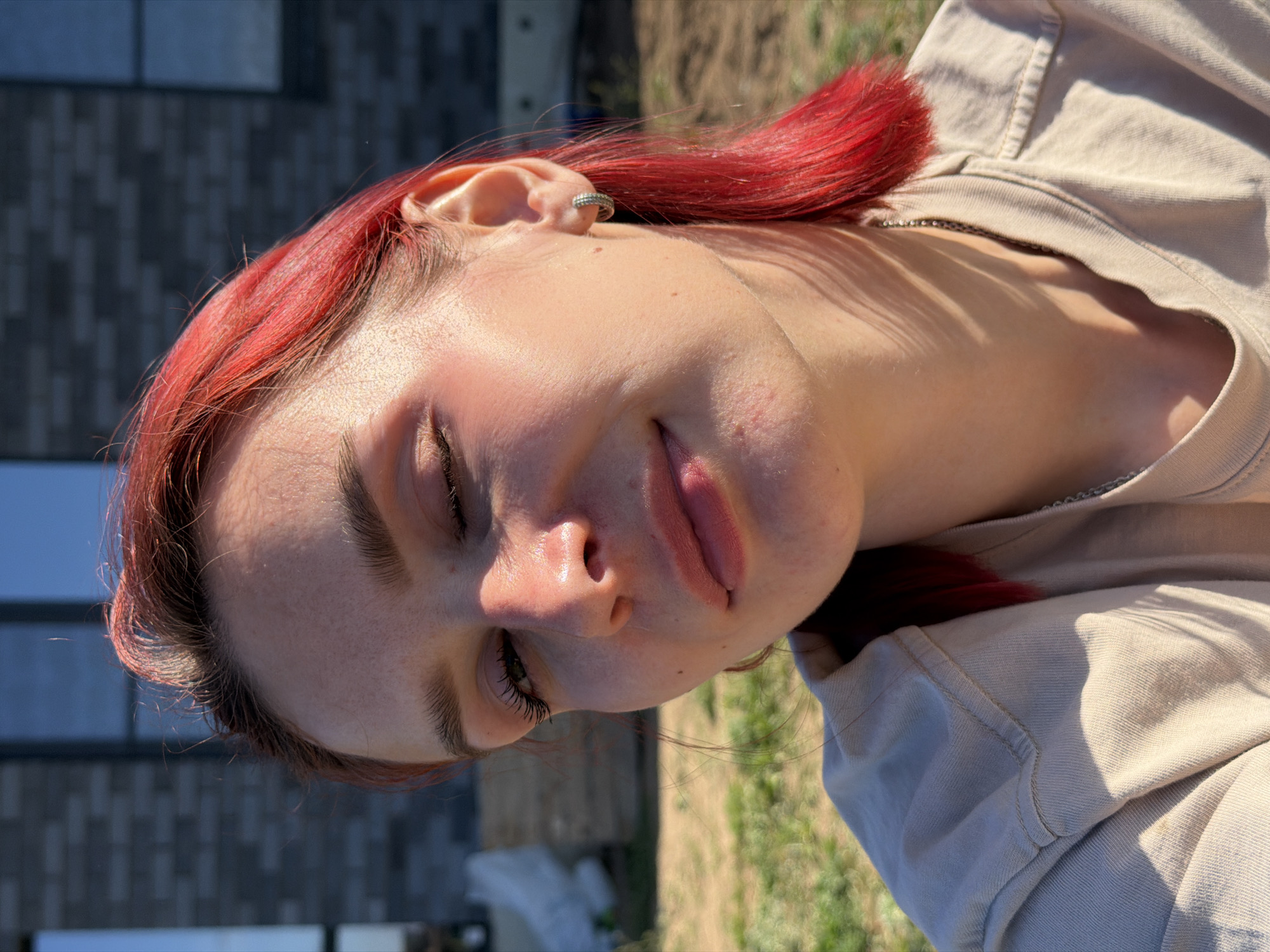 A close-up, slightly off-center, outdoor portrait of a young woman with red hair, light skin, and eyes closed. She is wearing a light-colored top, possibly a sweatshirt or jacket. She has an earring in her left ear. The background includes a building with a brick facade and some greenery, potentially grass.
While it's not possible to definitively pinpoint the location within Chaikovsky, Russia based solely on this image, the presence of brick buildings and vegetation suggests an urban or suburban area.