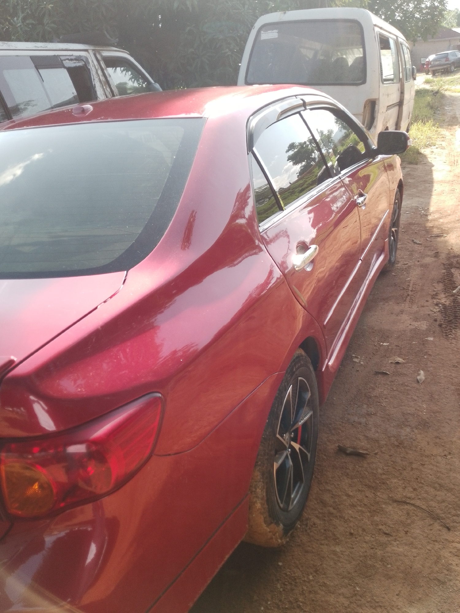 This media file displays a side view of a red sedan parked on a dirt road. The car's rear taillight and wheel are visible, along with a portion of its side panel and door handle. The car appears to be parked outdoors, with a white van and another vehicle, possibly an SUV, in the background. Trees and foliage frame the scene, suggesting a rural or semi-rural setting. The lighting indicates it is daytime, with the sun casting shadows and highlighting the car's reflective surface. The dirt road is uneven and appears somewhat muddy in places, hinting at recent rainfall or the general condition of the unpaved road. There is no text visible in the image.