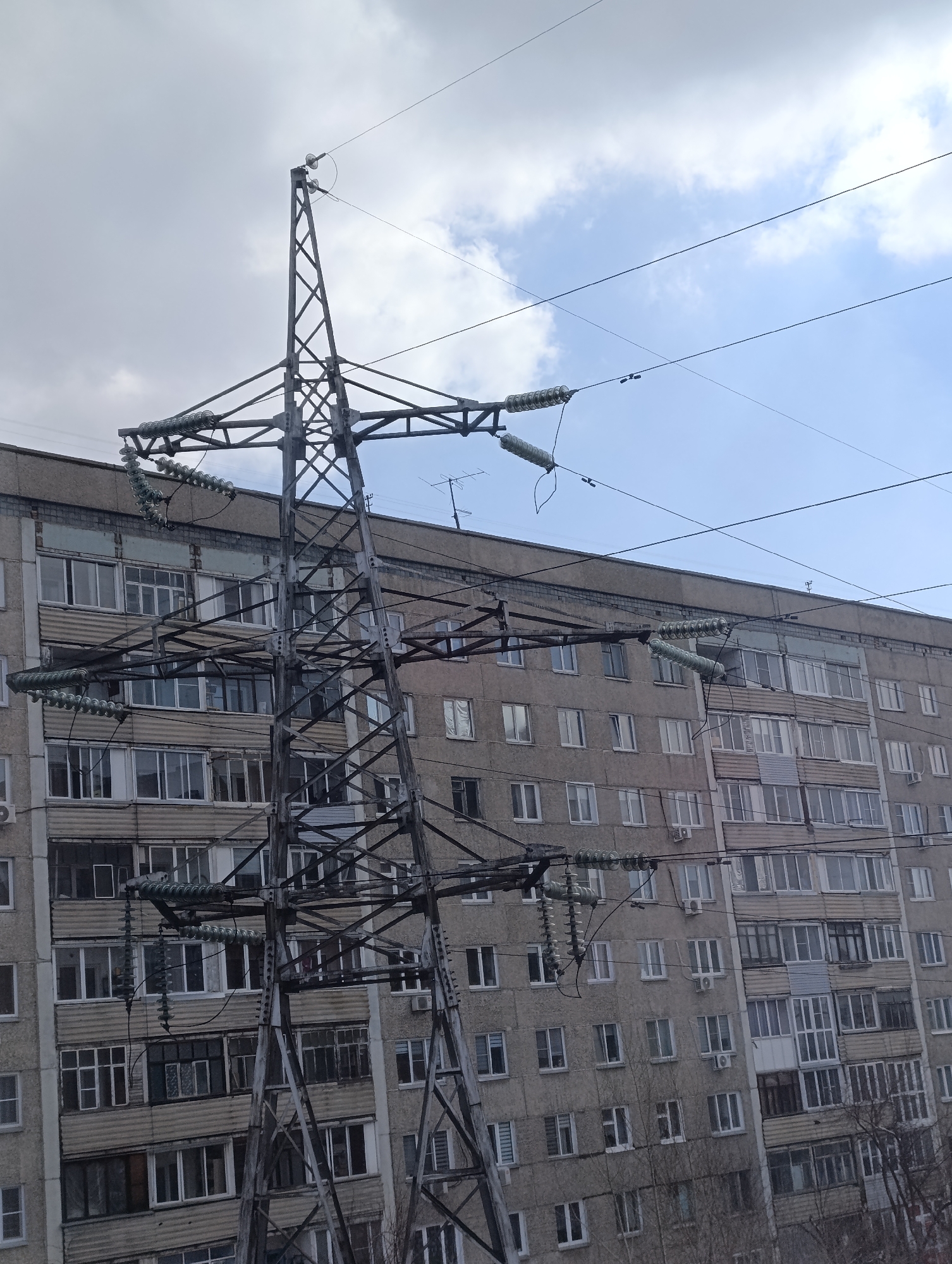 A large, grey metal lattice electricity pylon dominates the foreground and midground of the image, showcasing its intricate structural design. Multiple high-voltage power lines extend from the pylon, secured by long strings of light green or turquoise disc-shaped insulators. These lines stretch across the frame, some against the sky and others in front of the building in the background.
Behind the prominent pylon, a multi-story, grey concrete residential building fills the majority of the background. Its facade is densely packed with numerous windows and balconies, some of which appear enclosed or modified. Several white air conditioning units are affixed to the exterior walls below windows. An antenna system is visible on the building's roof. The architectural style of the building is characteristic of mass-produced housing found in Krasnoyarsk, Russia, and other post-Soviet cities.
The sky above presents a mixed weather condition, with large areas of overcast grey clouds alongside patches of bright blue sky dotted with white clouds, indicating daytime. The overall palette of the scene is muted, consisting mainly of greys from the pylon, building, and clouds. Bare tree branches are subtly visible in the lower right corner, suggesting a colder season.
The image captures a static urban or suburban scene in Krasnoyarsk, Russia, highlighting the integration of large-scale electrical infrastructure within a residential environment. No people or specific activities are depicted. There is no visible text in the image.