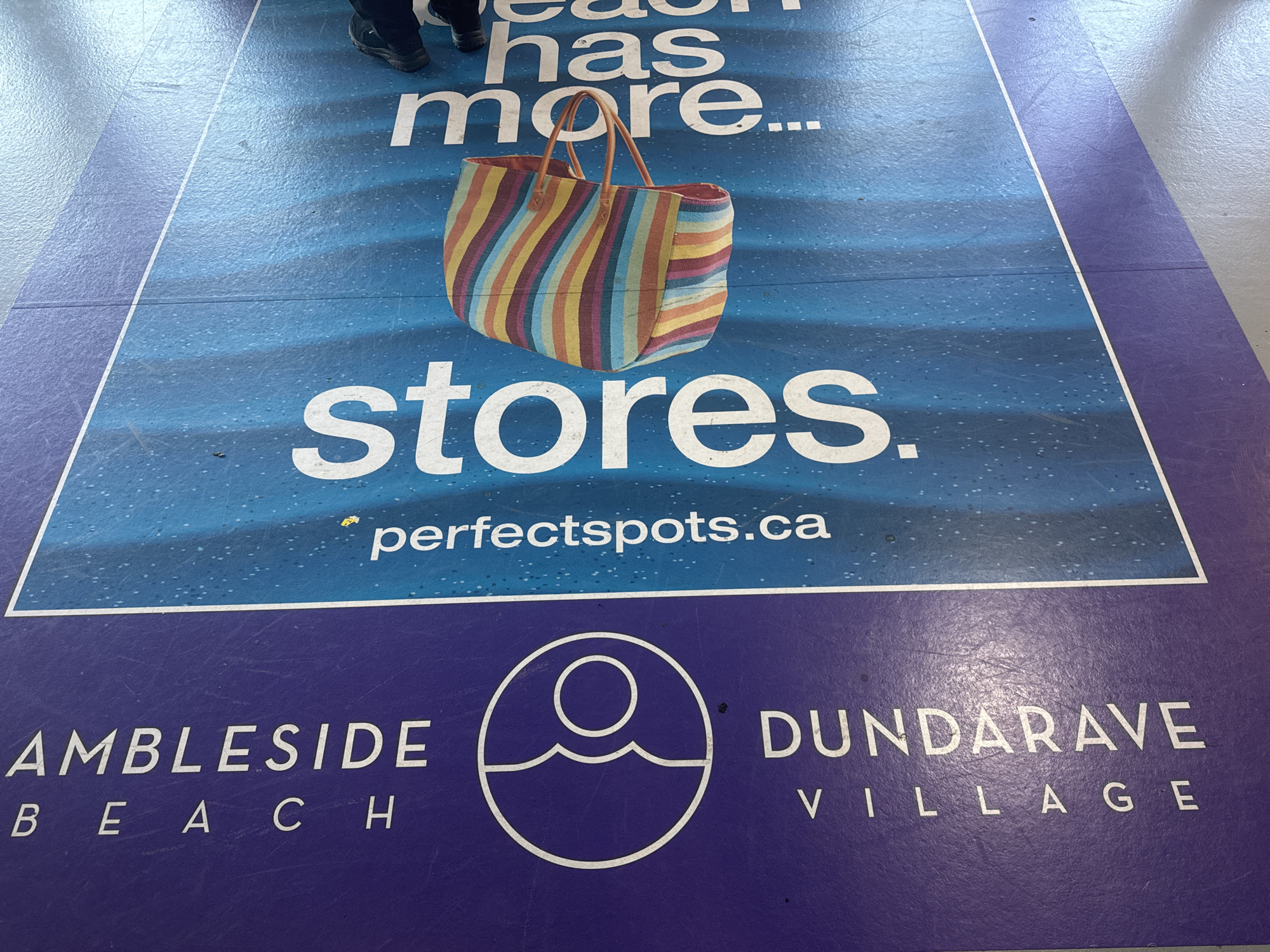 The image depicts a promotional floor graphic likely located in a transit station or mall in Vancouver, Canada. The graphic is divided into two main sections. The top section has a blue background with an image of a striped beach bag, text reading "beach has more... stores." and the website "perfectspots.ca". The lower section, on a purple background, has the text "AMBLESIDE BEACH" on the left and "DUNDARAVE VILLAGE" on the right, with a round graphic of a sun over a wave in the center. At the top of the image is a pair of feet wearing black shoes.