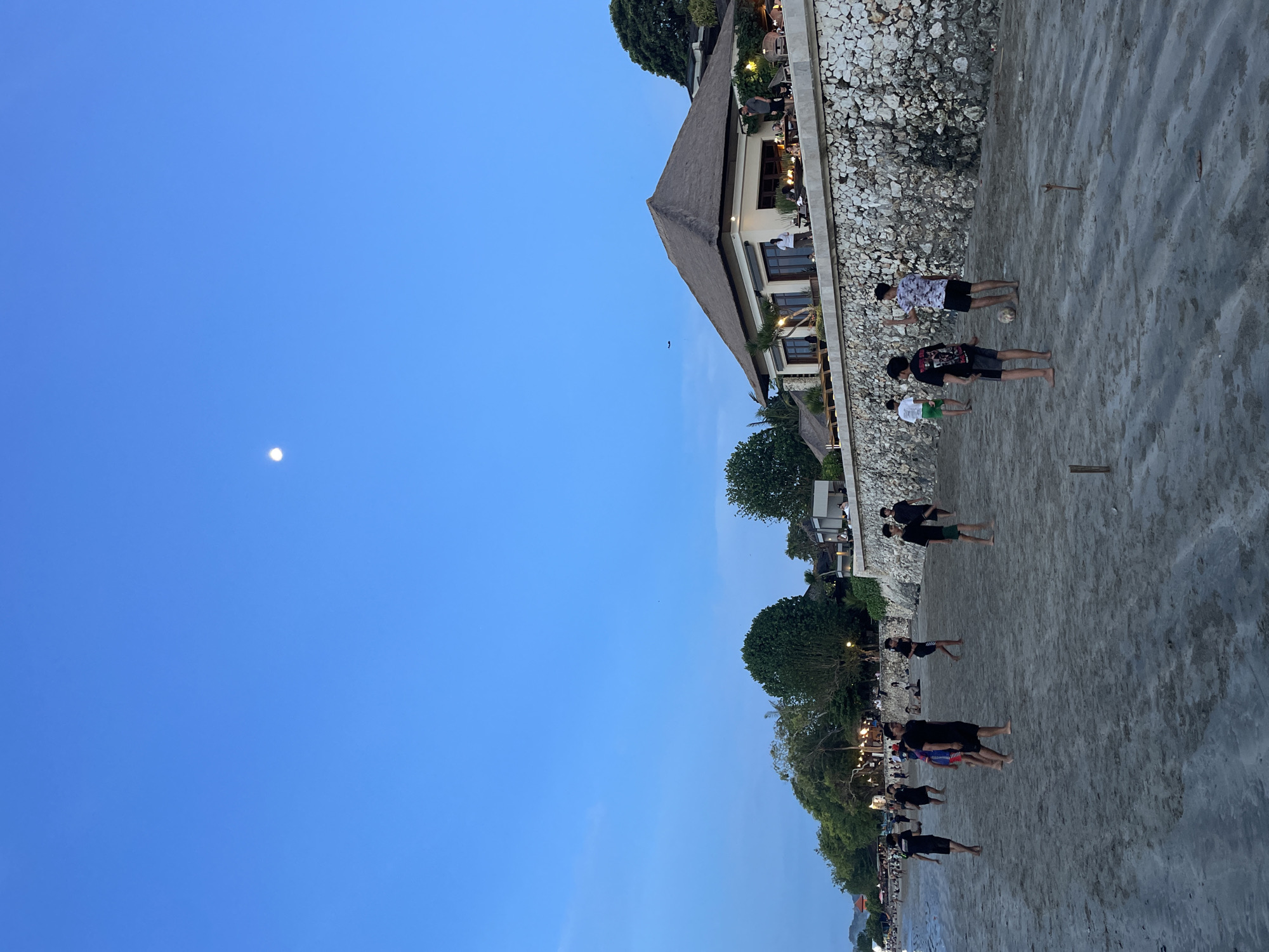 The image captures an evening scene on a sandy beach adjacent to a resort-like establishment. Numerous people, primarily young adults and men, are gathered on the beach, most of them standing in small groups or spread out, dressed casually in shorts and t-shirts. Some appear to be barefoot. A white ball, possibly for soccer or volleyball, rests on the sand near one group, suggesting recreational activity.
In the background, an ornate multi-story building with a traditional thatched roof and light-colored walls stands elevated above the beach, separated by a rugged stone retaining wall. Artificial lights are visible within the building and on its terraces, where other patrons can be seen. Lush green trees and foliage are scattered around the building and along the coastline, adding to the tropical ambiance.
The time of day is early evening or twilight, indicated by the clear, deep blue sky and the prominent, bright full moon visible in the upper left. Despite the moon's brightness, the sky retains a vibrant blue hue, suggesting it's not yet fully dark. The weather appears clear and calm, without any visible clouds. The setting, with its distinctive architecture and beach activities, is consistent with a tropical resort location like Kuta Selatan, Indonesia. No discernible text is visible in the image.
