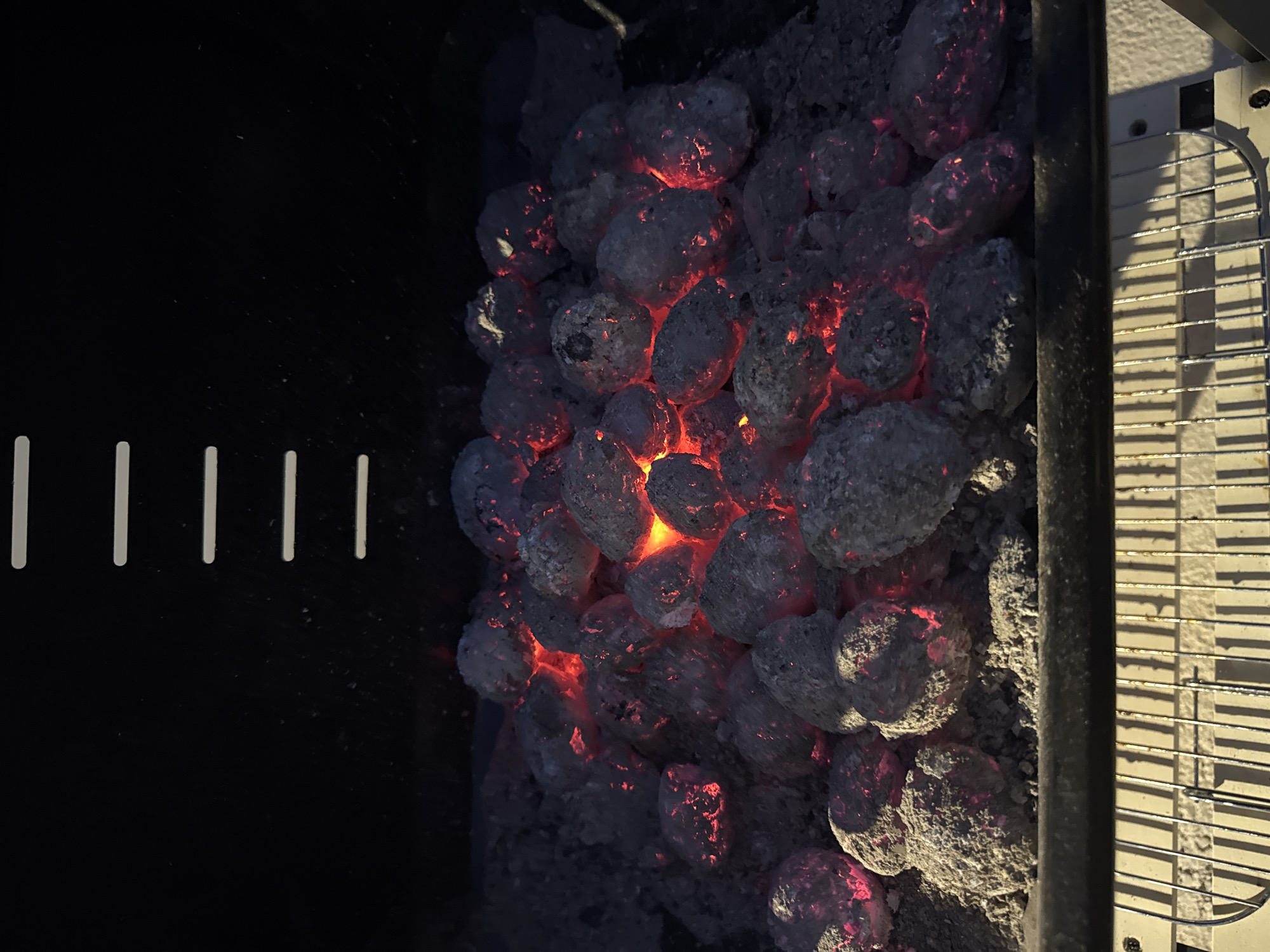 This is a close-up, high-angle shot of glowing charcoal briquettes inside a grill. The briquettes are piled in the center of the grill's firebox, with some embers radiating a bright orange-red light. The charcoal is covered in a layer of ash. The grill grates are visible at the bottom of the image. The scene is dark, with the only light source being the glowing coals. There are no people or animals in the image. The location is a grill, which is typically found in outdoor spaces such as backyards, patios, or parks.