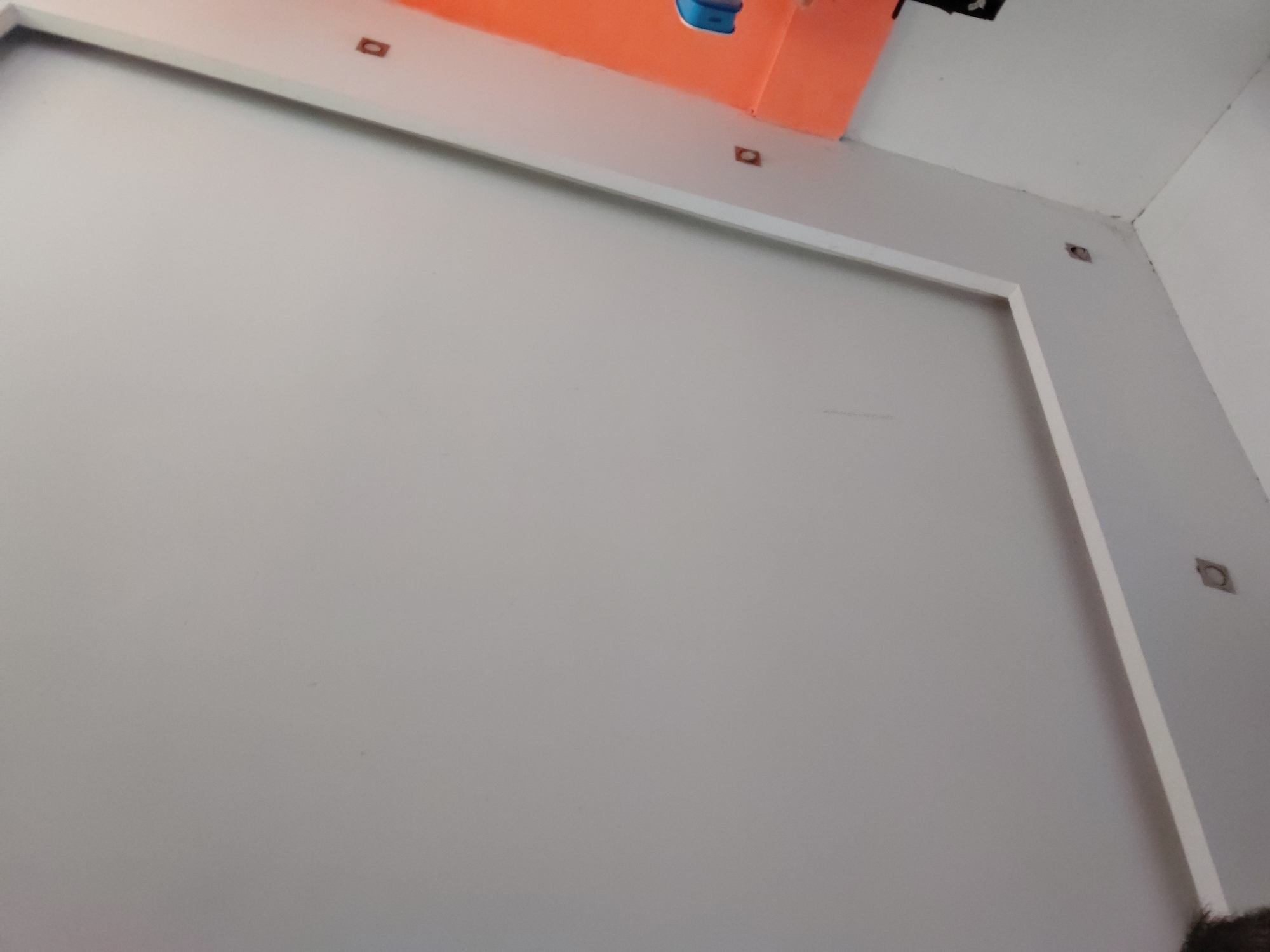 The image displays an indoor scene, likely a room or part of a building, located in Santa Ana, Venezuela. The central focus is a large, plain light grey wall. This wall is partially outlined by a clean white frame or border, creating a rectangular inset.
Along the top edge of this white frame, and extending onto the adjacent white wall and ceiling in the upper right corner, are several small, dark, square-shaped cutouts or holes. These openings suggest preparation for the installation of light fixtures, sensors, or other decorative elements.
In the upper left corner, a bright orange surface is visible, possibly part of a ceiling panel, soffit, or an architectural feature. A small blue object, which appears to be a fixture or device, is situated on this orange surface. Further to the right in the upper corner, a small black object is also discernible.
The photograph is taken from a low angle, looking upwards, with a small portion of what appears to be a person's head or hair visible at the very bottom right edge of the frame. The lighting is bright and even, indicating an indoor environment, possibly with artificial light. The scene depicts a static interior space, possibly under construction or renovation, with a clean and minimalist aesthetic. No discernible activity or text is present in the image.