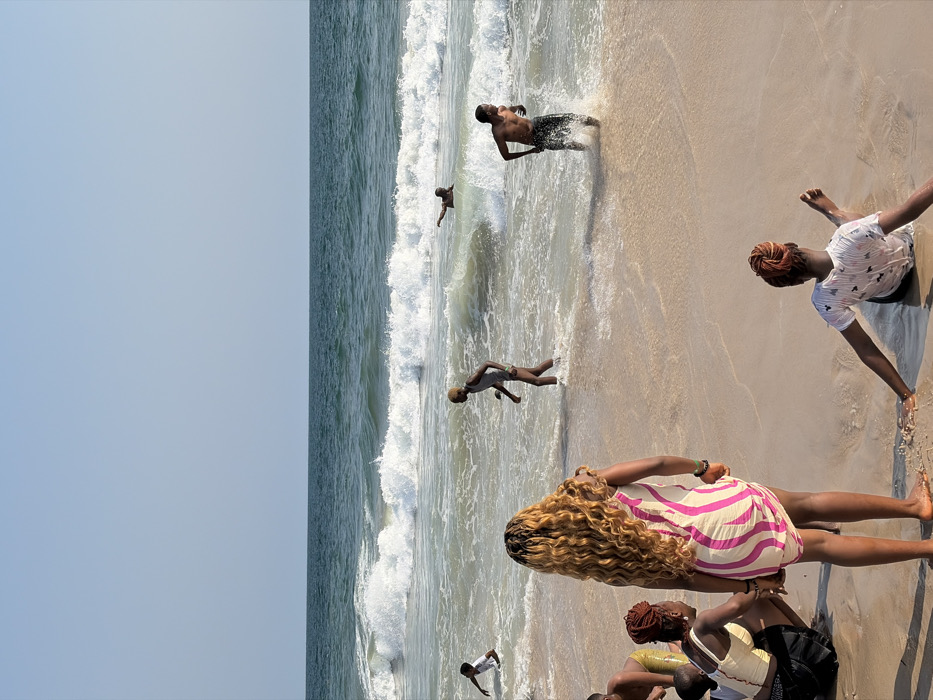 The scene depicts a sunny beach in Mopo Onijebu, Nigeria, with a clear blue sky meeting the ocean horizon. Approximately nine individuals are present, engaged in various activities. In the immediate foreground, a person with long, blonde curly hair stands facing the ocean, wearing a light-colored dress with vertical pink stripes. To their left, another person sits on the sand with dark hair, partially visible. To the right, a person with red-orange hair tied in a bun sits in the shallow wet sand, facing away, wearing a white patterned t-shirt, with arms extended.
In the midground, within the breaking waves, a bare-chested male with dark skin is depicted running towards the shore, splashing water. To his left, another individual with light-colored short hair, wearing light swimwear, walks further into the water. Further out in the ocean, a person is partially submerged, extending an arm. Another person, a smaller figure in a white shirt, is visible in the shallow water to the far left. The ocean displays moderate white-capped waves rolling towards the sandy shore. The environment is characterized by light-colored sand and bright natural light.