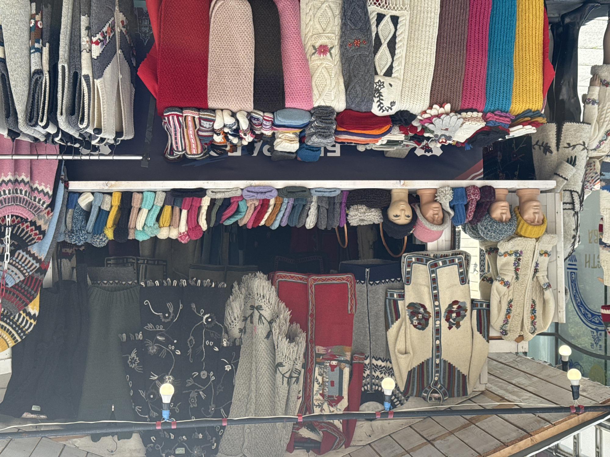 This is a close-up, upside-down, eye-level shot of a market stall selling knitted goods. The stall features a variety of sweaters, scarves, hats, and gloves in a wide range of colors and patterns, including traditional folk designs. Many of the items are handmade and appear to be made of wool.
In the foreground, several mannequins with heads are displayed, wearing knitted hats and some with visible faces. The background shows more shelves filled with knitted items. The overall impression is of a vibrant and colorful market offering warm, handcrafted clothing.
The image appears to have been taken during the daytime, with artificial lighting from a few bulbs visible near the bottom. There are no clear indicators of the weather or specific emotions. The location is implied to be Belgrade, Serbia, based on the metadata, though no explicit text in the image confirms this. There is some faint text visible on the back wall of the stall, but it is illegible.