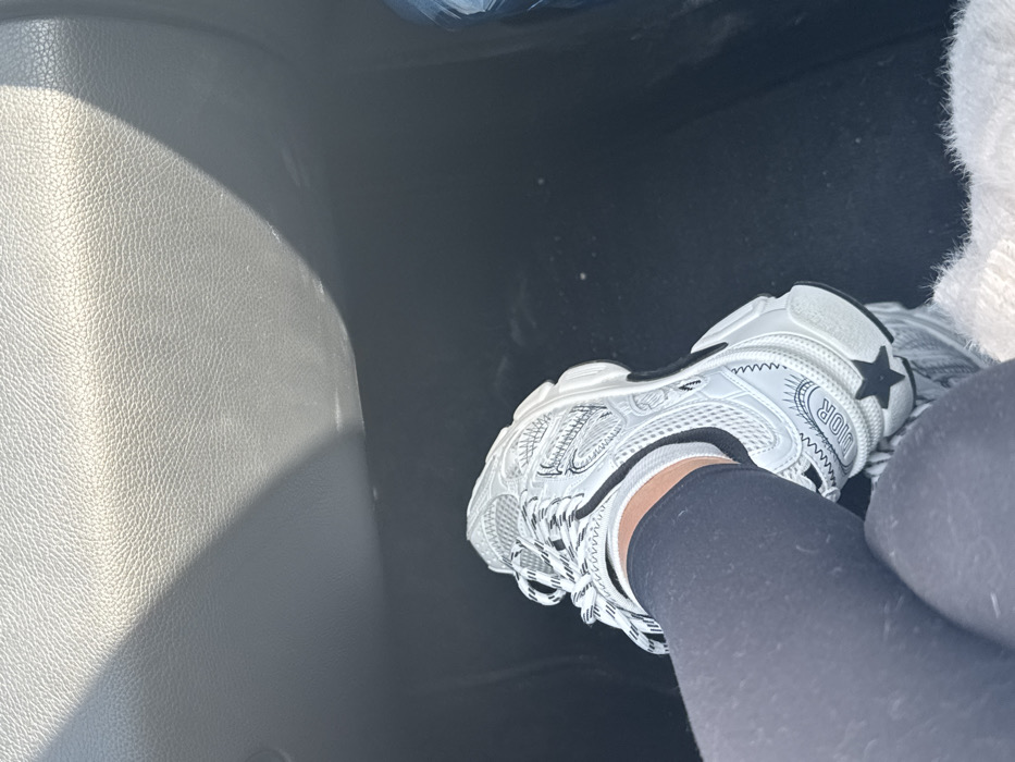 The image displays a close-up, downward-angled view of a person's lower leg and foot positioned within the passenger compartment of a vehicle. The individual is wearing black form-fitting trousers or leggings, a white sock, and a white, chunky sneaker. The sneaker is clearly branded "DIOR" on the heel, featuring black and white striped laces and a black star emblem on its side. A portion of a white, fluffy textile, possibly a garment or blanket, is visible in the lower right foreground. The immediate environment includes a dark, textured grey vehicle dashboard or interior panel in the upper portion, illuminated by sunlight casting a distinct shadow. The vehicle's floor is dark-colored. No specific visual elements within the image confirm the requested location of Sofia, Bulgaria; the scene is consistent with a generic vehicle interior.