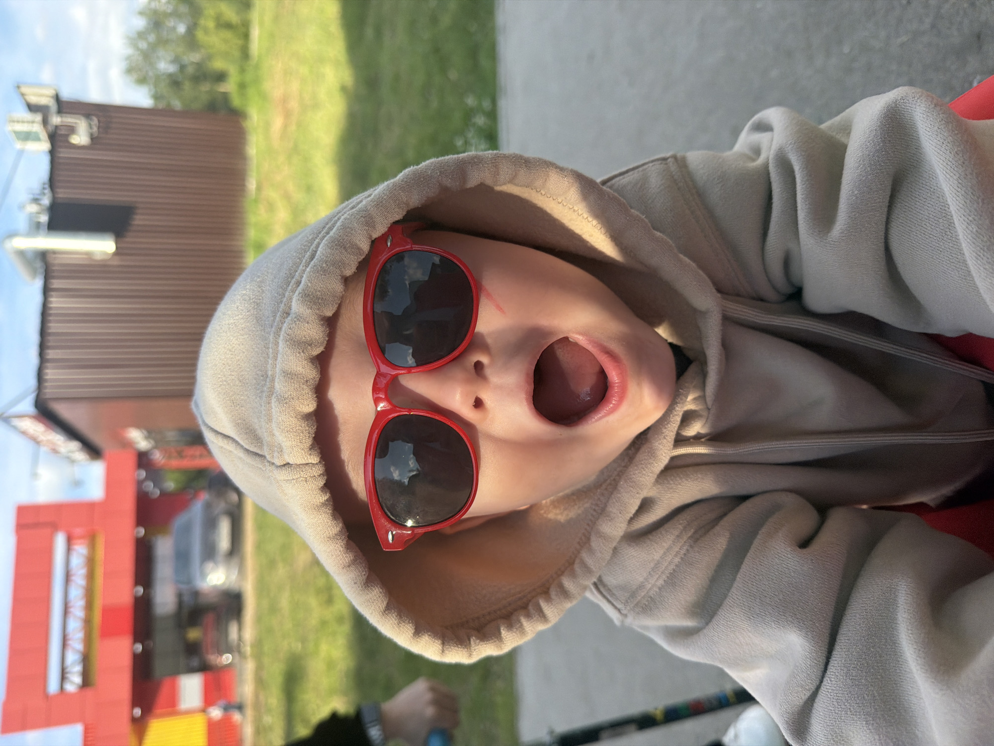 The media file is a vertical, close-up, selfie-style photograph of a young child wearing a beige hooded sweatshirt and red sunglasses. The child's mouth is open, and their tongue is visible. In the background, out of focus, is a brown building with metal siding, a bright red and yellow structure that appears to be part of a playground or commercial building, and some greenery. A dark-colored car is also visible in the background. The lighting suggests it is daytime, likely outdoors in fair weather, with shadows indicating sunlight. The overall impression is of a playful, candid moment. The location is identified as Chaikovsky, Russia. There is no visible text in the image.