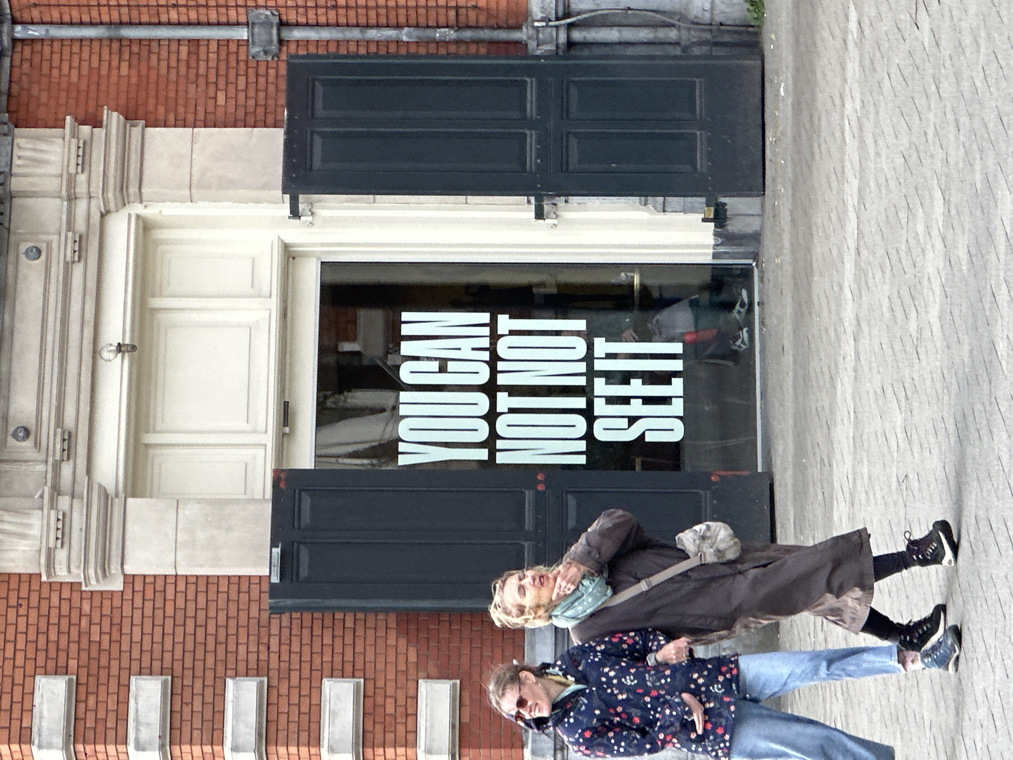 The image shows a building facade on a brick building in Amsterdam, Netherlands, with a sign on a window that reads "YOU CAN NOT NOT SEE IT" in vertically stacked white block letters. Two women are standing on the sidewalk in front of the building. The woman in the foreground wears a blue floral jacket, blue jeans, and sunglasses. The woman behind her wears a long brown coat and a scarf. The facade consists of brick and stone, with a large door frame and decorative elements. The doorway has dark doors on top and bottom and a glass window in the center. The sidewalk is paved with grey stones. A drainpipe runs vertically along the brick wall, and a conduit extends horizontally above the facade.