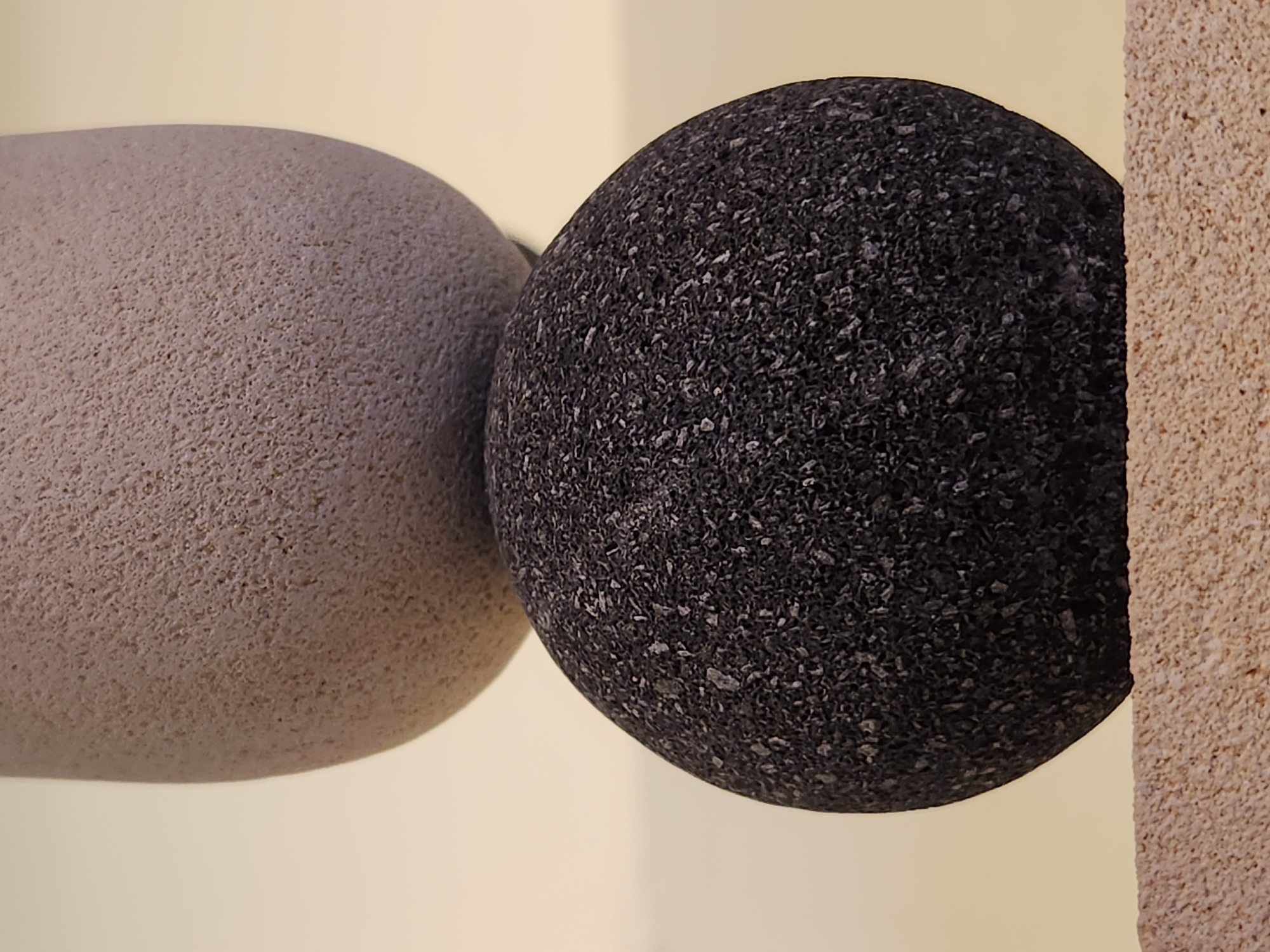 The image shows a close-up of two spherical objects placed next to a textured, vertical surface. The object on the left is larger and appears to be a light gray, possibly stone or concrete sphere with a granular texture. The object on the right is smaller, a dark gray or black sphere, also with a granular texture, and it is positioned slightly in front of and to the right of the lighter sphere. A portion of a beige, rough-textured vertical surface is visible on the far right of the image, partially obscuring the dark sphere. The background is a plain, light yellow, suggesting an indoor setting or a diffused outdoor light. There is no discernable activity taking place, and the scene is static. The lighting appears even, with no strong shadows, indicating it might be daylight but not direct sunlight. No text is visible in the image. The overall impression is one of abstract composition focusing on texture and form. The setting is not identifiable beyond the general interior or studio context.
