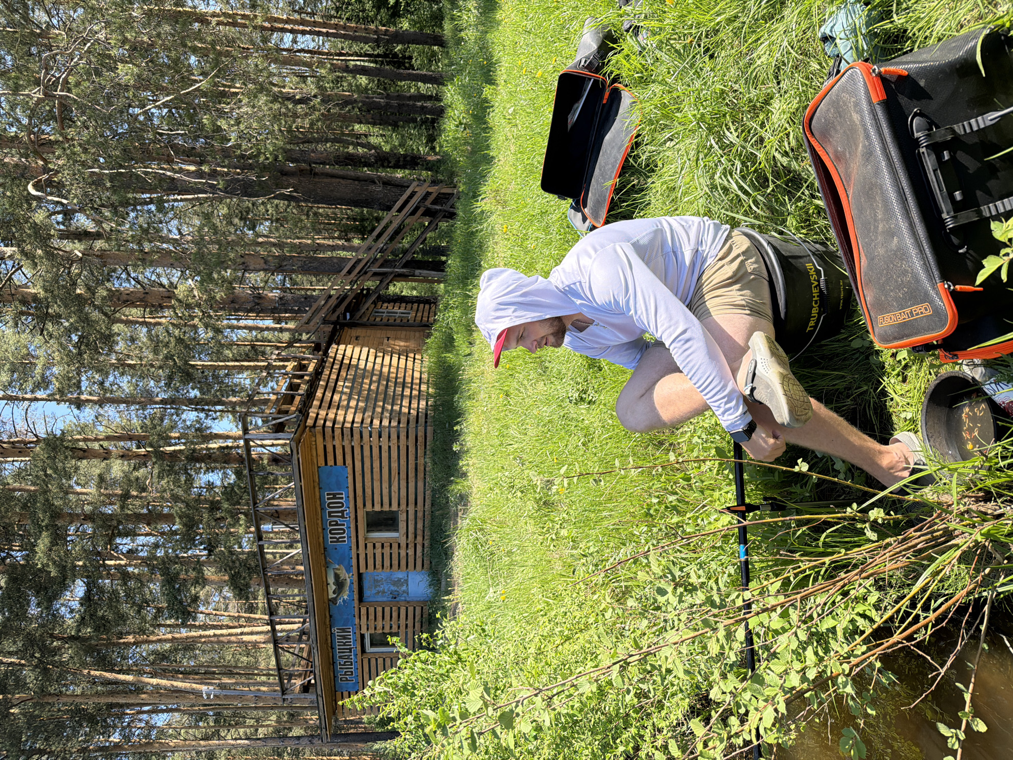 The image shows a man in a white hooded sweatshirt and khaki shorts, kneeling on grass next to a body of water. He appears to be adjusting something near a fishing rod. A black bucket and black bags with orange trim are situated nearby on the grass. In the background is a wooden building with a blue sign displaying text in Russian, identifying it as "Кордон Рыбацкий" (Fisherman's Cordon), possibly a fishing lodge or outpost. The location appears to be Прикамский, Russia. Tall pine trees surround the area, indicating a wooded environment.