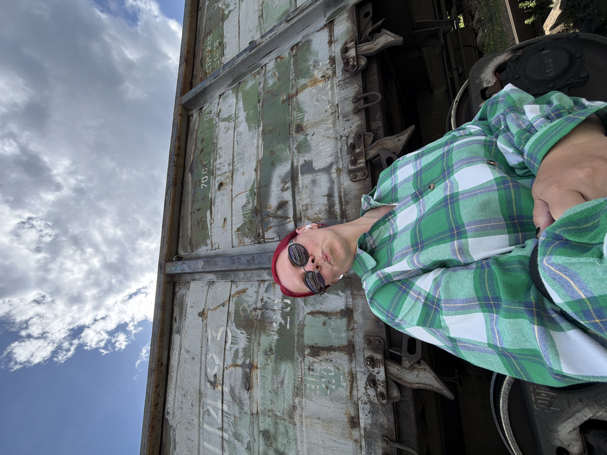 The media file shows a person wearing a green and white plaid shirt and sunglasses, resting their head against a weathered, green shipping container. The person has red hair and is making a kissing face. The shipping container is old, with visible rust and peeling paint, and bears some markings and text, including "70 OT" and Cyrillic lettering. The scene is set outdoors, with a blue sky and dramatic clouds visible in the background, suggesting it is daytime. The person appears relaxed and playful. The overall setting suggests an industrial or transportation-related area, possibly near train tracks or a port, given the presence of the shipping container and what appears to be a train wheel in the foreground. The provided location context indicates Chaikovsky, Russia.