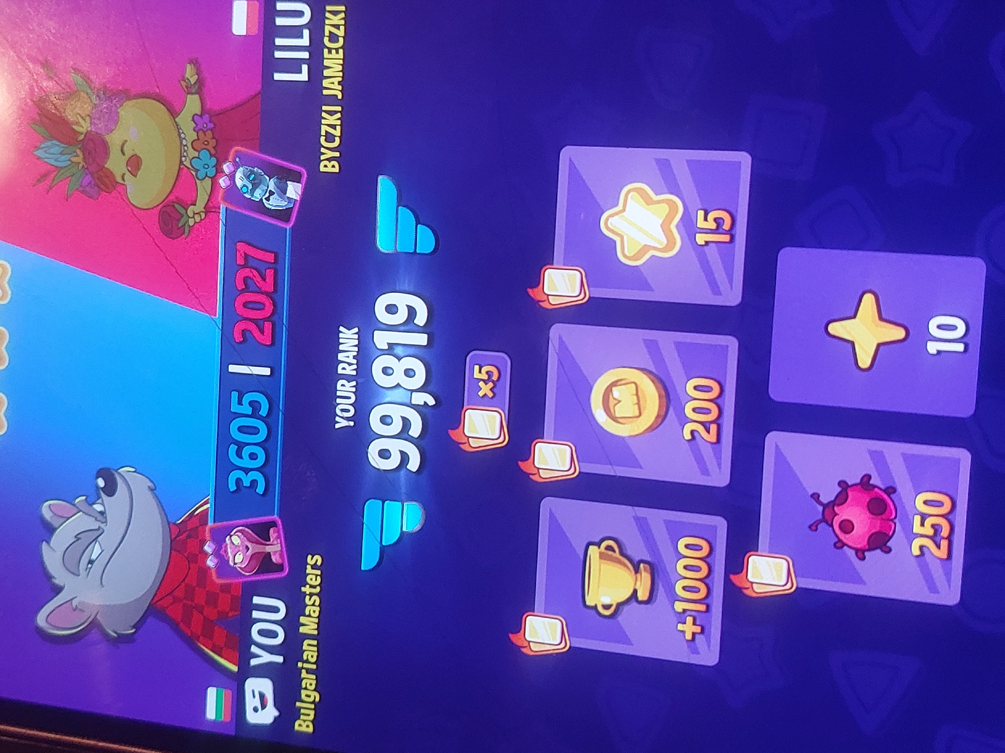 The image shows a screen capture from a mobile game. On the left side, there are two cartoon characters: one is a wolf wearing a red checkered vest, labeled "YOU" with a Bulgarian flag; the other is a yellow hippopotamus-like figure with a flower crown, labeled "LILU" with a Polish flag. The player "YOU" has a score of 3605, while "LILU" has a score of 2027. The "YOUR RANK" displayed is 99,819. To the right, there are purple cards with gold icons, showing rewards: 15 stars, 200 coins, 1000 points (trophy icon), 10 stars, and 250 ladybug icons. The rewards include a x5 bonus. The text "Bulgarian Masters" is visible under "YOU" implying the game is being played in Bulgaria. However, the image does not specify that it is in Tsarevo.