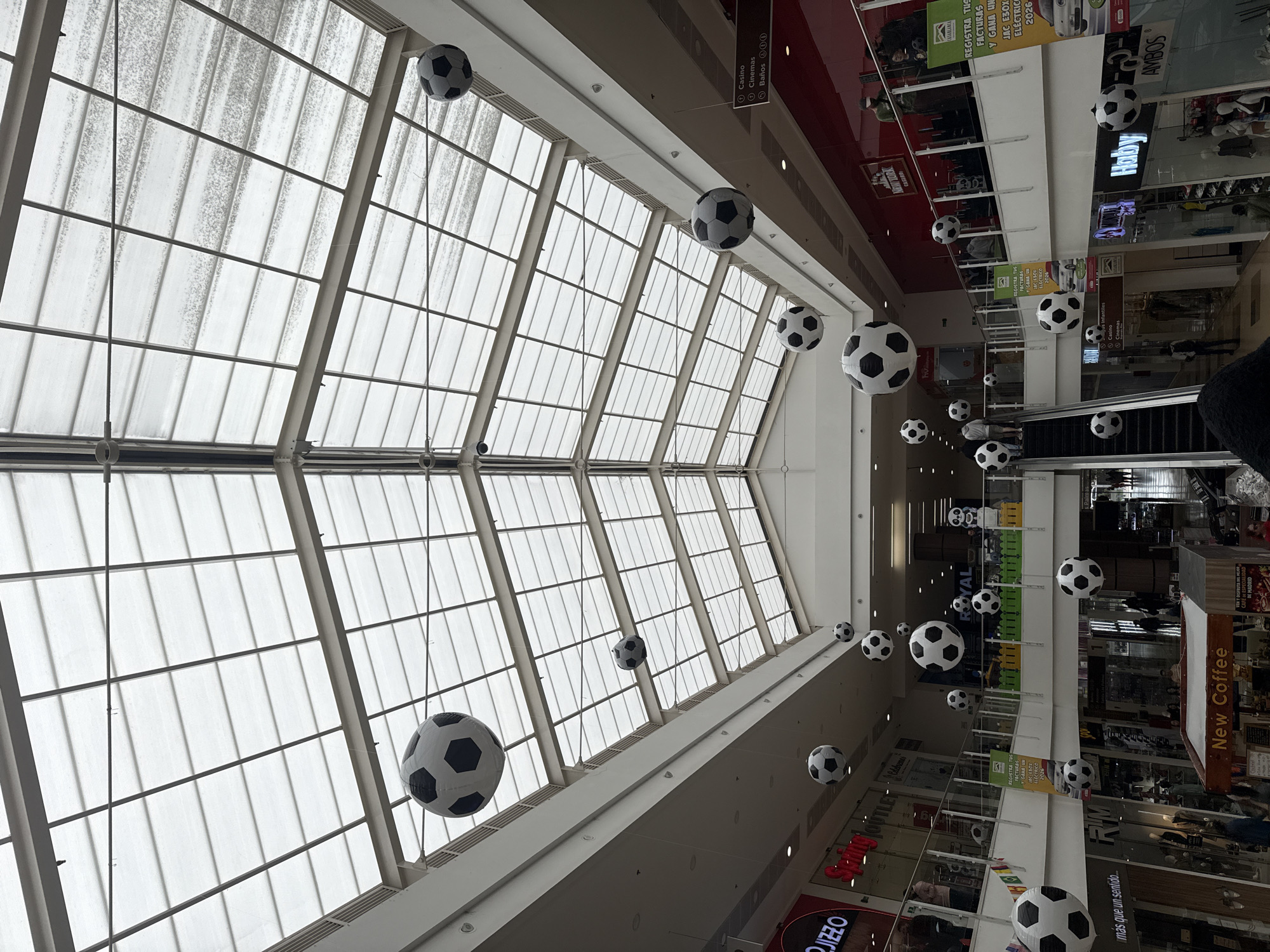 The image depicts the interior of a multi-story shopping mall located in Madrid, Colombia.
A prominent gabled skylight, composed of numerous translucent rectangular panels set within a light-colored metal framework, forms the ceiling, allowing diffuse natural light into the space. Throughout the central atrium, numerous black-and-white soccer balls of varying sizes are suspended from the ceiling at different elevations, serving as the primary decorative theme.
The mall features at least three visible levels, connected by escalators and framed by white railings with glass inserts. On the lower left, an escalator descends, while another ascends, partially visible on the right side of the frame. Various retail storefronts line the upper levels, displaying names such as "JIZZO," "Spring OUTLET," "ROYAL," "New York Cereals," "Habby," and "Ambos." A "New Coffee" stand with a wooden counter is visible on the ground floor.
Informational signage in Spanish includes directions to "Casino," "Cinemas," and "Baños" (bathrooms) on the upper right wall. On the left side, a large yellow banner advertises a promotion to "REGISTRA TUS FACTURAS Y GANA UN JAC E30X ELÉCTRICO 2026" (Register your invoices and win a JAC E30X Electric 2026). Small decorative flags with colors resembling the Colombian flag are draped over a railing on an upper level on the left. A large white and black panda statue stands on the upper right level, near the "ROYAL" sign.
Approximately a dozen individuals are scattered throughout the mall, walking on different levels and escalators. Their actions are limited to general movement; no specific interactions or detailed activities are discernible. The overall environment is well-lit and presents as a typical contemporary shopping center.