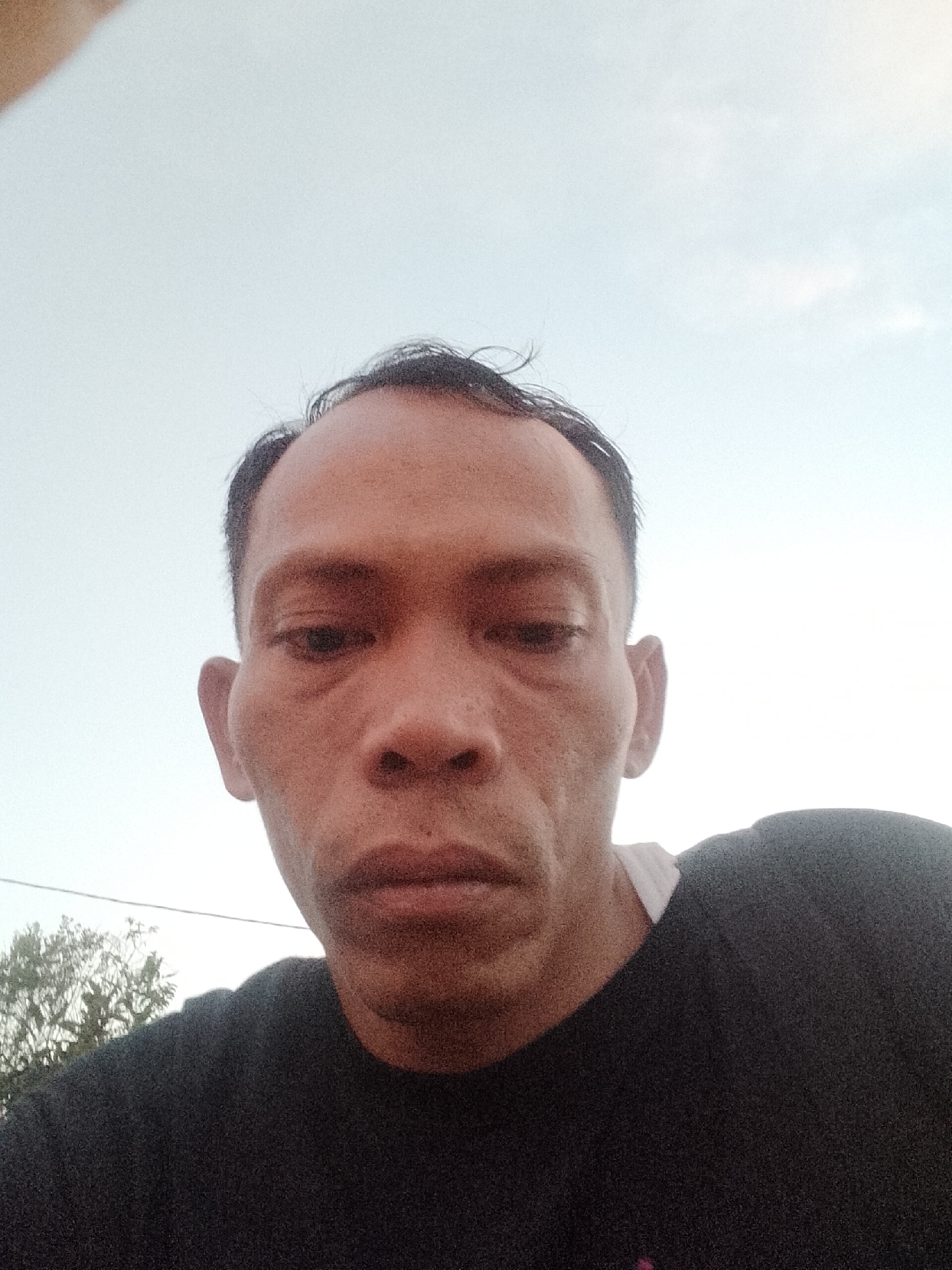 A selfie of a man in Muara Sungsang, Indonesia, shows his head and shoulders against a pale sky. He is wearing a black t-shirt with a white collar visible. The man has short, dark hair and appears to be looking down towards the camera. The lighting suggests it is daytime, possibly late afternoon or early morning, due to the soft, diffused light. The background is mostly sky with a hint of green foliage to the left, indicating an outdoor setting.