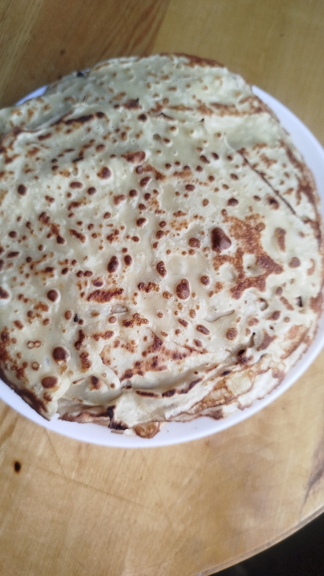 A stack of crepes is presented on a white plate, which rests on a wooden surface. The crepes are golden brown with darker spots, indicating they are cooked. The wooden surface appears to be a table with visible grain. The background is out of focus. No people are visible in the image. The scene is indoors.