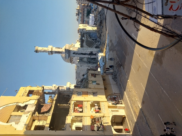 This image captures a street scene in Khan Yunis, Palestinian Territory, during daylight hours. The prominent feature on the left is a multi-story, light-colored building exhibiting severe structural damage, with large sections of its facade and interior walls missing, exposing multiple rooms. Laundry is visible hanging on some balconies of the intact portions of the building, suggesting continued habitation. On the ground level, two individuals, appearing to be children, walk on a sidewalk in the lower left corner.
The central area features an asphalt road with visible white lane markings, partially covered by shadows. Along the road's edges and extending into the midground are substantial accumulations of construction debris and rubble. A group of approximately five individuals is clustered near a large pile of rubble in the midground, while a single individual walks further down the street in the distance.
In the background, a large mosque with a prominent dome and a tall minaret stands, also displaying considerable structural damage to its main building. Extensive piles of rubble and the remains of other demolished buildings surround the mosque. Further in the distance, additional urban structures are visible under a clear, bright sky. A portion of a dark, circular wire structure and a white box with Arabic text are visible in the lower right foreground, partially obscuring the view.