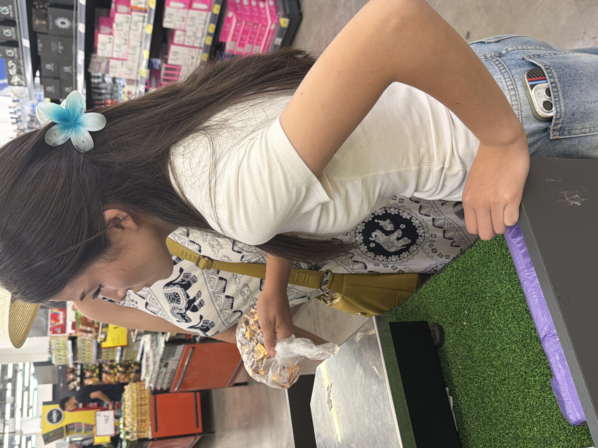 The image shows a young woman in a white t-shirt and denim shorts standing in a grocery store. She is holding a plastic bag of candy and is about to place it on a scale. Her hair is dark and long, adorned with a blue and white flower clip. Behind her, other shoppers and store shelves are visible. The environment appears to be a brightly lit supermarket aisle with various products displayed on shelves. The woman's hand is resting on a black counter. A roll of purple bags is visible at the bottom of the frame. Her phone is in her back pocket.