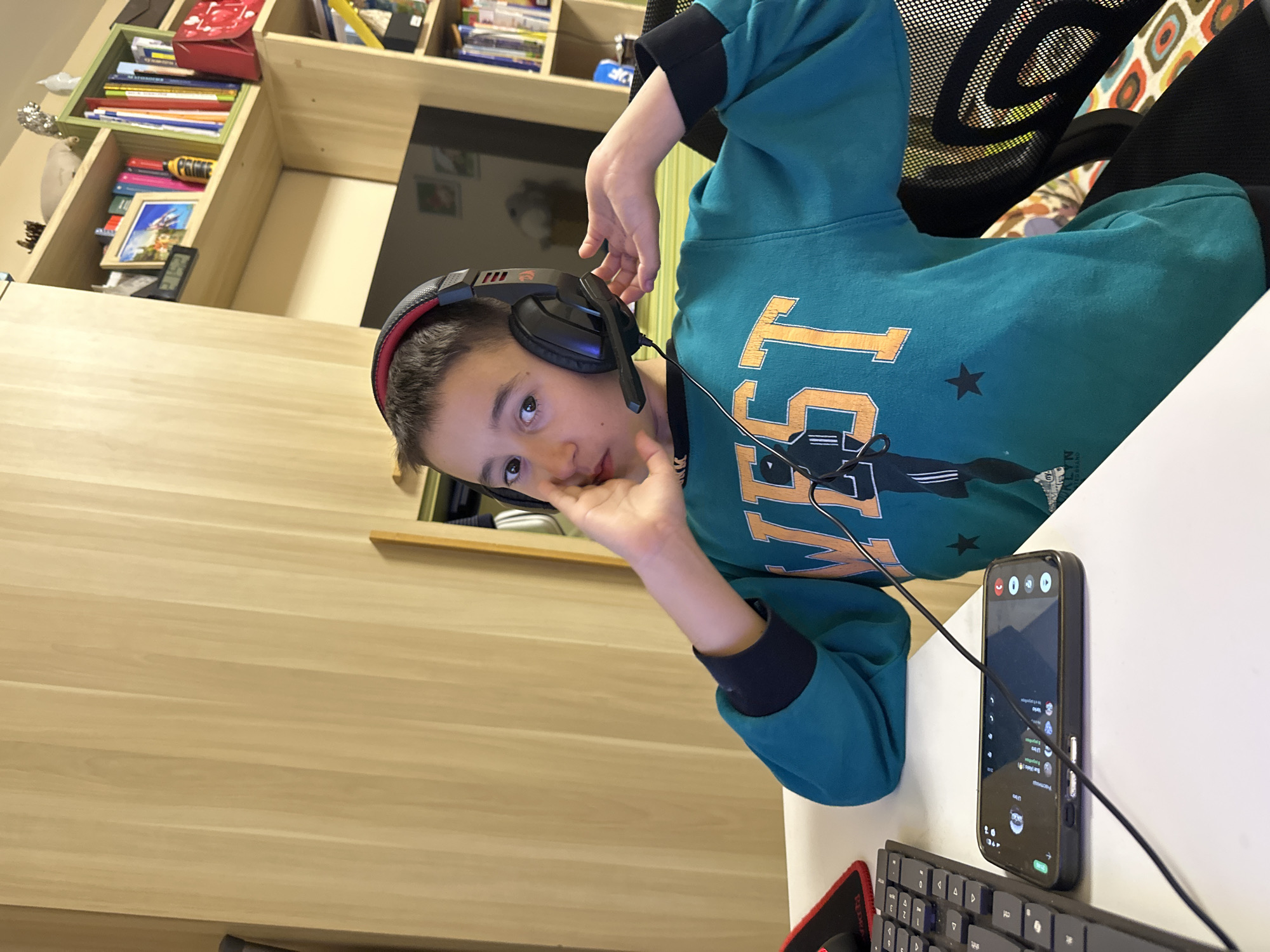 A young boy, likely a pre-teen, is captured indoors, possibly in a home study or gaming area in Plovdiv, Bulgaria. The image, oriented vertically, shows the boy looking directly at the camera with a neutral expression. He has short dark hair and is wearing a teal-colored long-sleeved hooded sweatshirt with black cuffs. The sweatshirt features large orange text that reads "WEST," a black star, and other graphic elements.
He is wearing black gaming headphones with red accents and a microphone boom positioned near his mouth, suggesting he is engaged in an activity requiring audio communication. One hand is raised, lightly touching his chin or mouth, while the other rests on the headphone cup.
The boy is seated at a white desk. On the desk, a black smartphone stands upright, displaying a messaging application interface, possibly Discord, with visible chat entries and a username "Lino." A portion of a black computer keyboard is also visible. In the background, a light wooden bookshelf is filled with various books, a red gift bag, and decorative items. A yellow object on one shelf has the word "PRIME" visible. A black mesh office chair is partially seen to his right, and a television screen or monitor is reflected in the distance. The scene is well-lit, indicating an indoor activity during daylight hours or under artificial lighting.