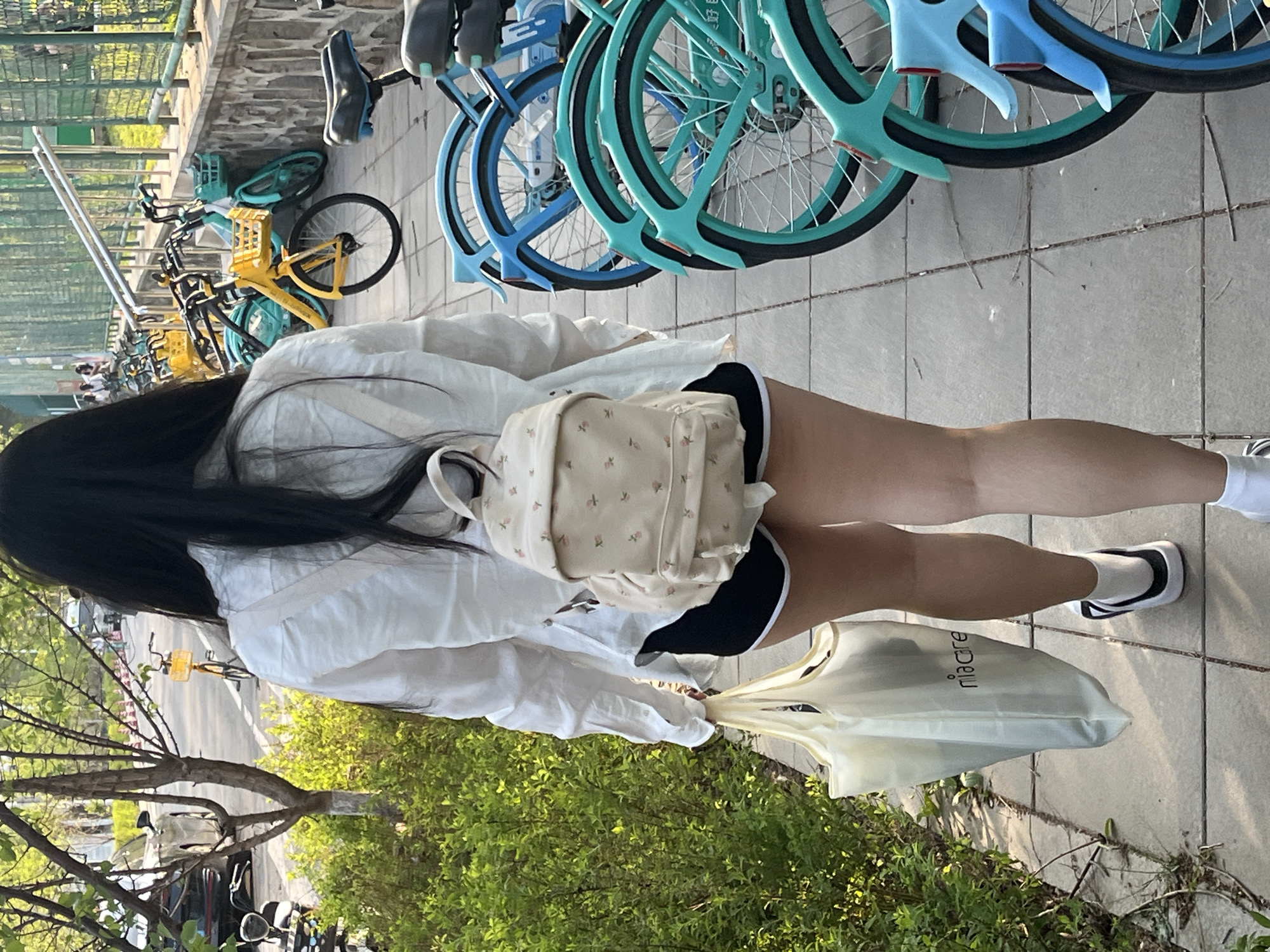 A medium shot depicts a young woman with long black hair, a white backpack with a floral pattern, a white button-down shirt, black shorts, and white socks with black accents, walking on a tiled sidewalk adjacent to parked shared bicycles in the city of Changchun, China. She carries a white plastic shopping bag labeled "niacare." To her left, a low hedge is present, and behind her, a line of shared bikes and racks is visible.