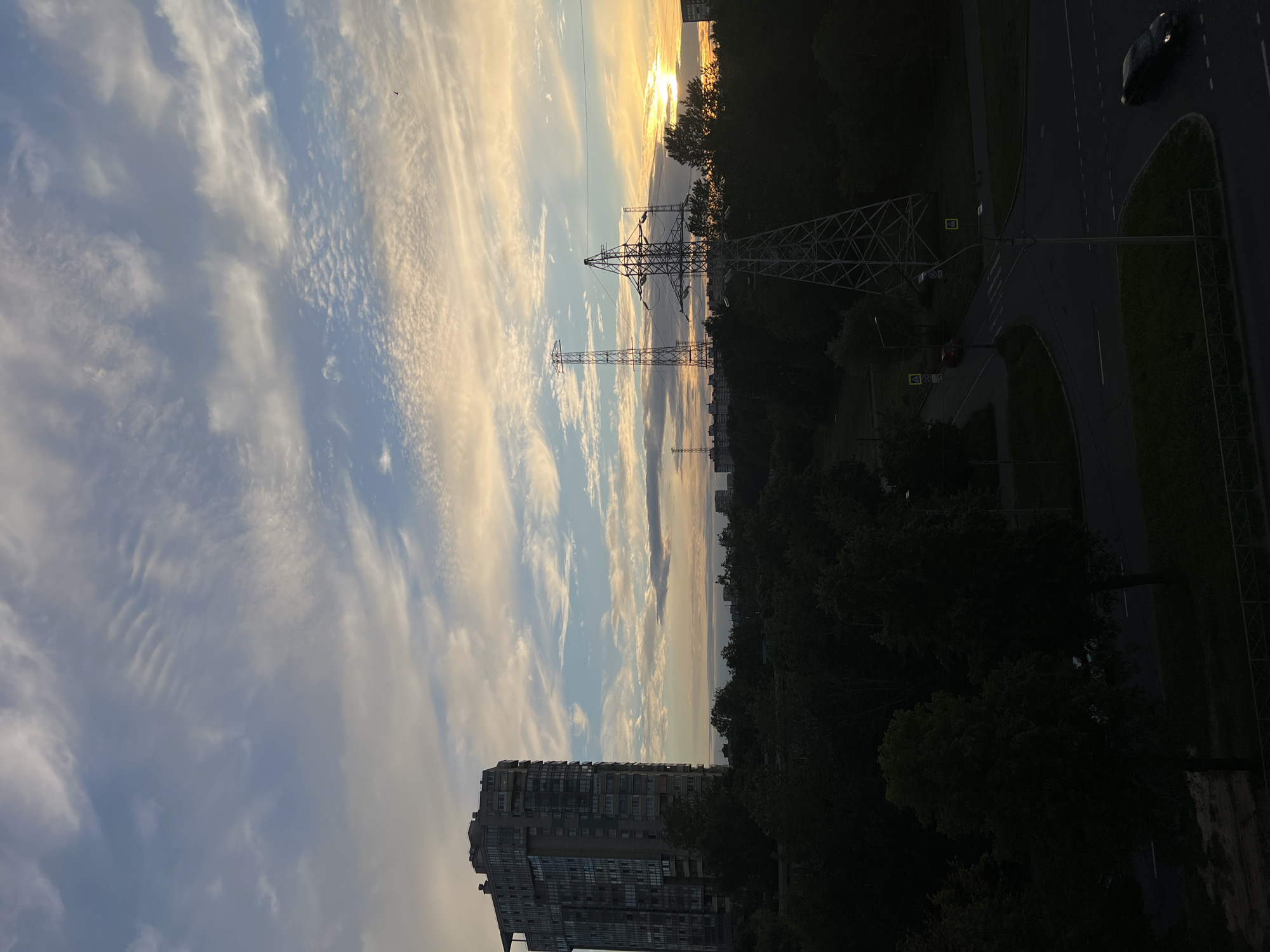 This is a vertical, outdoor, daytime photograph taken from an elevated viewpoint in Saint Petersburg, Russia. The scene captures a juxtaposition of urban infrastructure and natural elements under a dramatic sky.
Dominating the left side of the frame is a tall, modern apartment building, its facade a grid of windows and balconies. To the right, silhouetted against the bright sky, are two large electricity pylons. Beyond them, a dense canopy of dark green trees forms a natural barrier, obscuring much of the ground level. A paved road with lane markings is visible on the far right, with a dark-colored car driving on it. The road appears to be part of a larger urban intersection, with pedestrian crossings and road signs visible, though not clearly legible.
The sky is a prominent feature, filled with wispy, elongated clouds painted in shades of white, pale blue, and warm yellow and orange hues, indicating either sunrise or sunset. The sun itself is a bright, diffused light source near the horizon, peeking through the clouds and casting a warm glow over the scene. A small bird is visible in the sky on the left.
The overall mood is serene yet dynamic, with the grandeur of the sky contrasting with the ordered structure of the buildings and infrastructure. The time of day is clearly either early morning or late evening, suggested by the quality of light and the presence of the clouds.