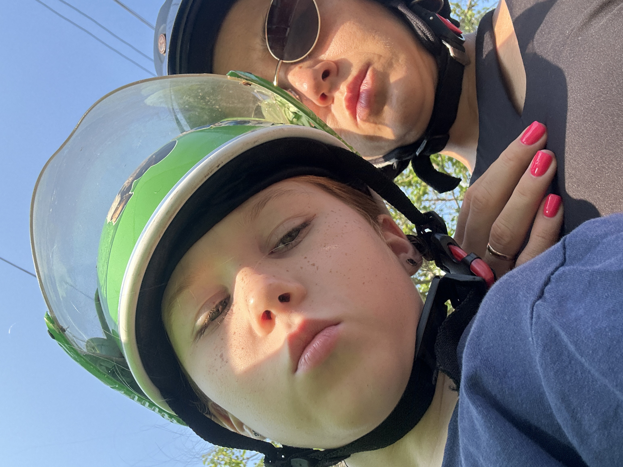 A low-angle close-up shot depicts two individuals wearing helmets. The person in the foreground has freckles and is wearing a green and white helmet with a clear visor. The person behind is wearing sunglasses and a black helmet, with one hand resting near the helmet strap of the person in front. Both appear to be wearing dark-colored clothing. The background reveals a blue sky and hints of green foliage. The location is Bac Nha Trang Ward, Vietnam.