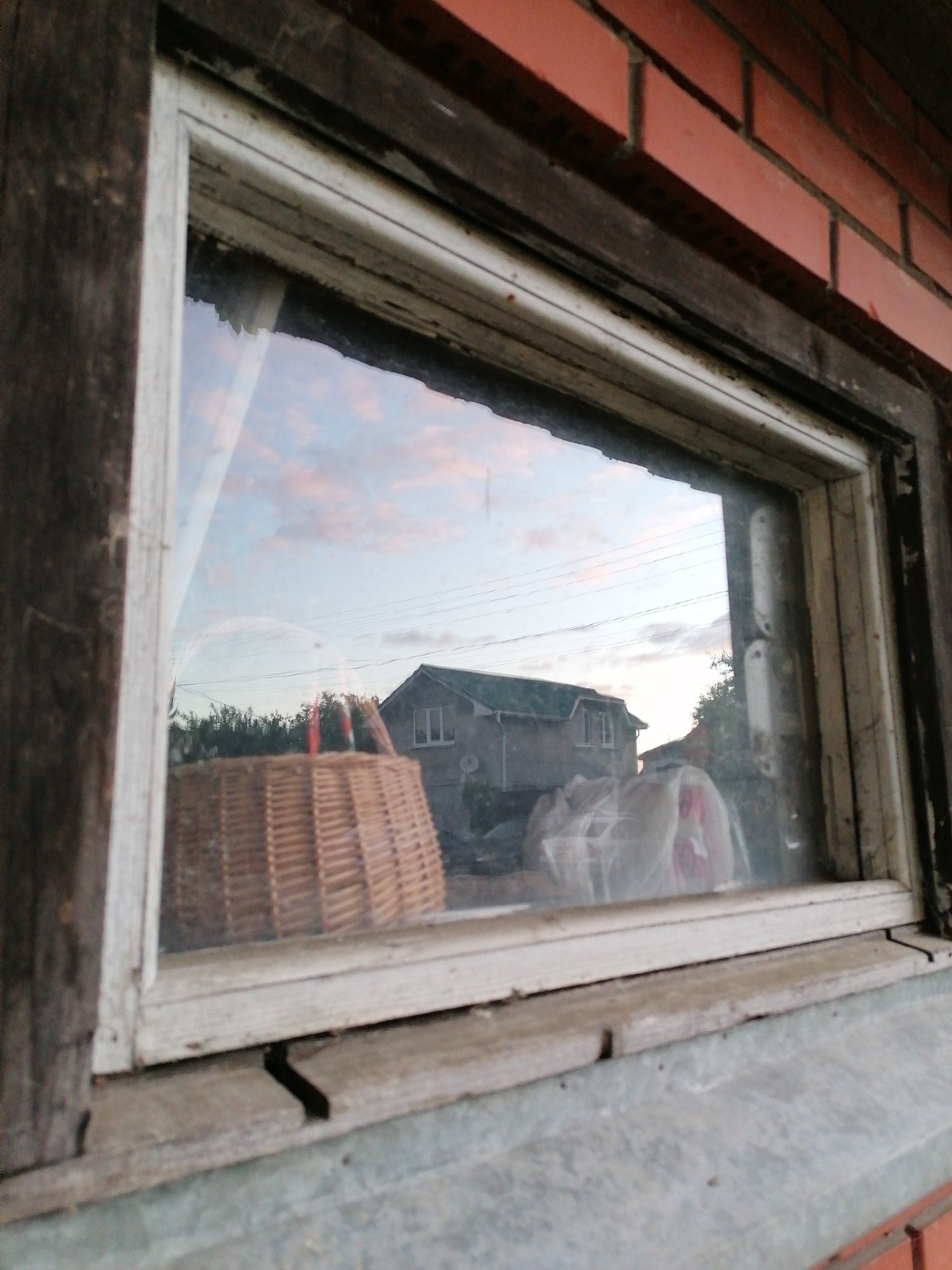 Here is a description of the image.
The image shows a view through a window, likely of an exterior scene. A wooden frame surrounds the window, parts of which are painted white while other sections are left as bare wood. The window pane reflects a view of a building across the way, a two-story structure with a green roof and white window frames. Wicker basket in the foreground. Some utility lines are visible in the sky above the building, and the sky itself is partly cloudy with a pinkish hue, possibly indicating dusk or dawn. The wall around the window is made of red brick. The foreground also includes a section of the window sill, which appears to be made of stone or concrete, and has some cracks and dirt on the surface.