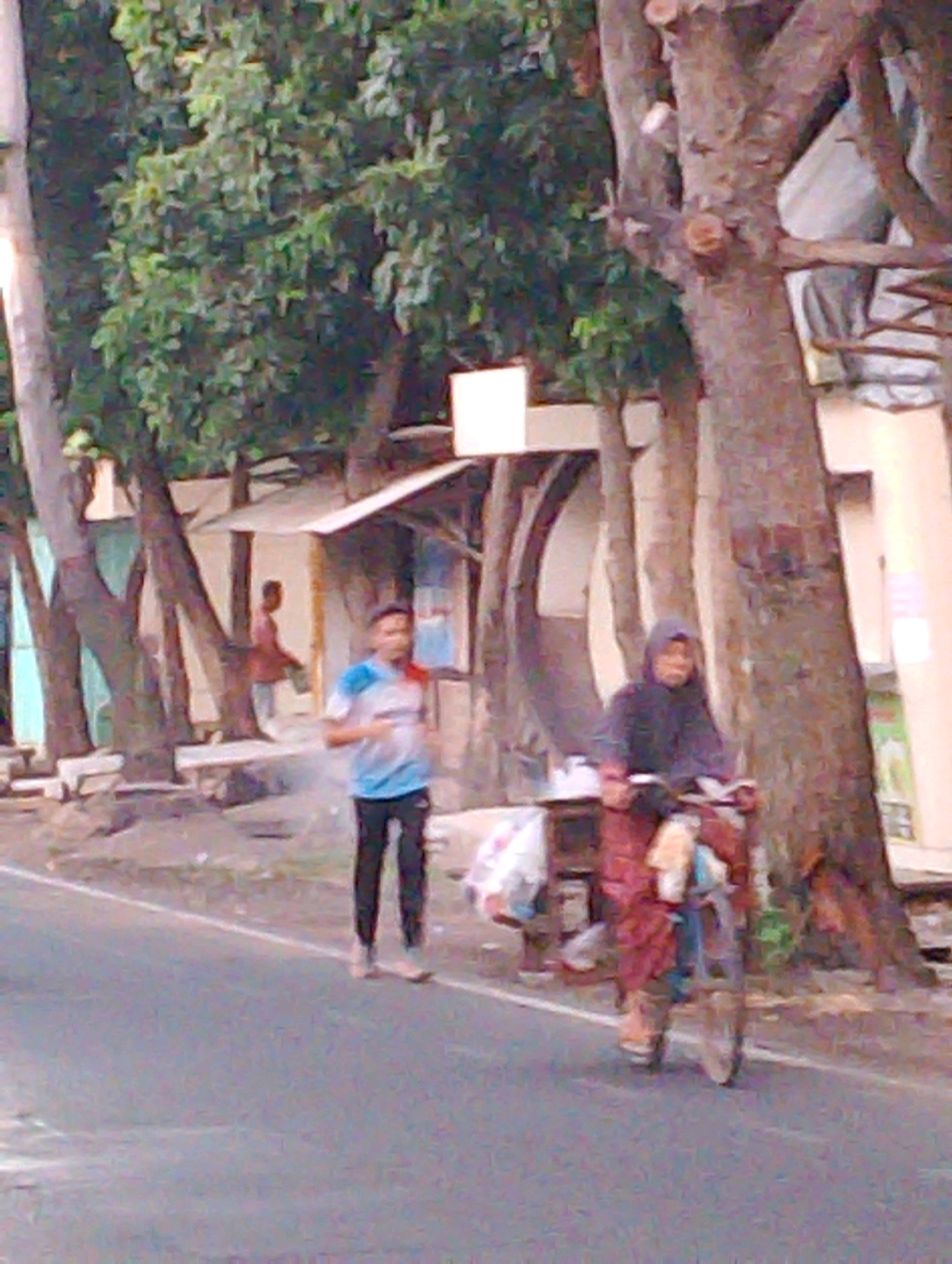 This image captures an outdoor scene with two people on a street. In the foreground, a person is riding a bicycle from right to left, wearing a dark-colored hooded garment and a red skirt or dress. The bicycle has a basket or rack on the back with a white and beige bag. To the left of the cyclist, another person is running from right to left, wearing a white t-shirt with blue and red accents and black pants. Behind the runners, a small wooden structure, possibly a stall or hut, is visible with a canopy. Several trees with rough bark line the background, some with pruned branches. The ground is paved with asphalt, and a white line marks the edge of the road. The lighting suggests it is daytime, possibly late afternoon. The overall environment appears to be a street in a tropical or subtropical region, likely a residential or commercial area.