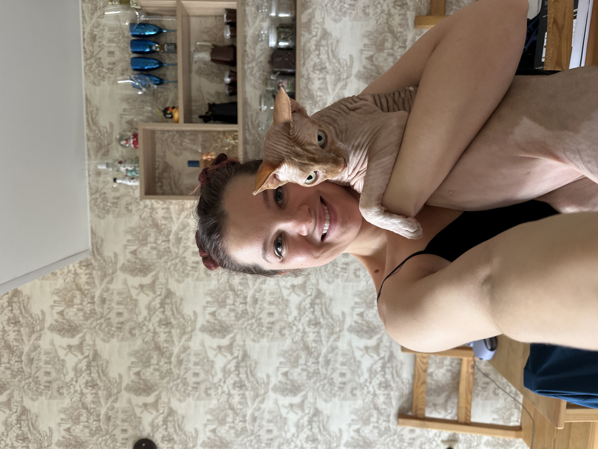 A woman holds a Sphynx cat. She is smiling and looks at the camera. The cat looks at the camera as well. Behind them are shelves with various items. The wallpaper is patterned. A partial view of a wooden chair can be seen. The location is in the city of Vladimir, Russia.