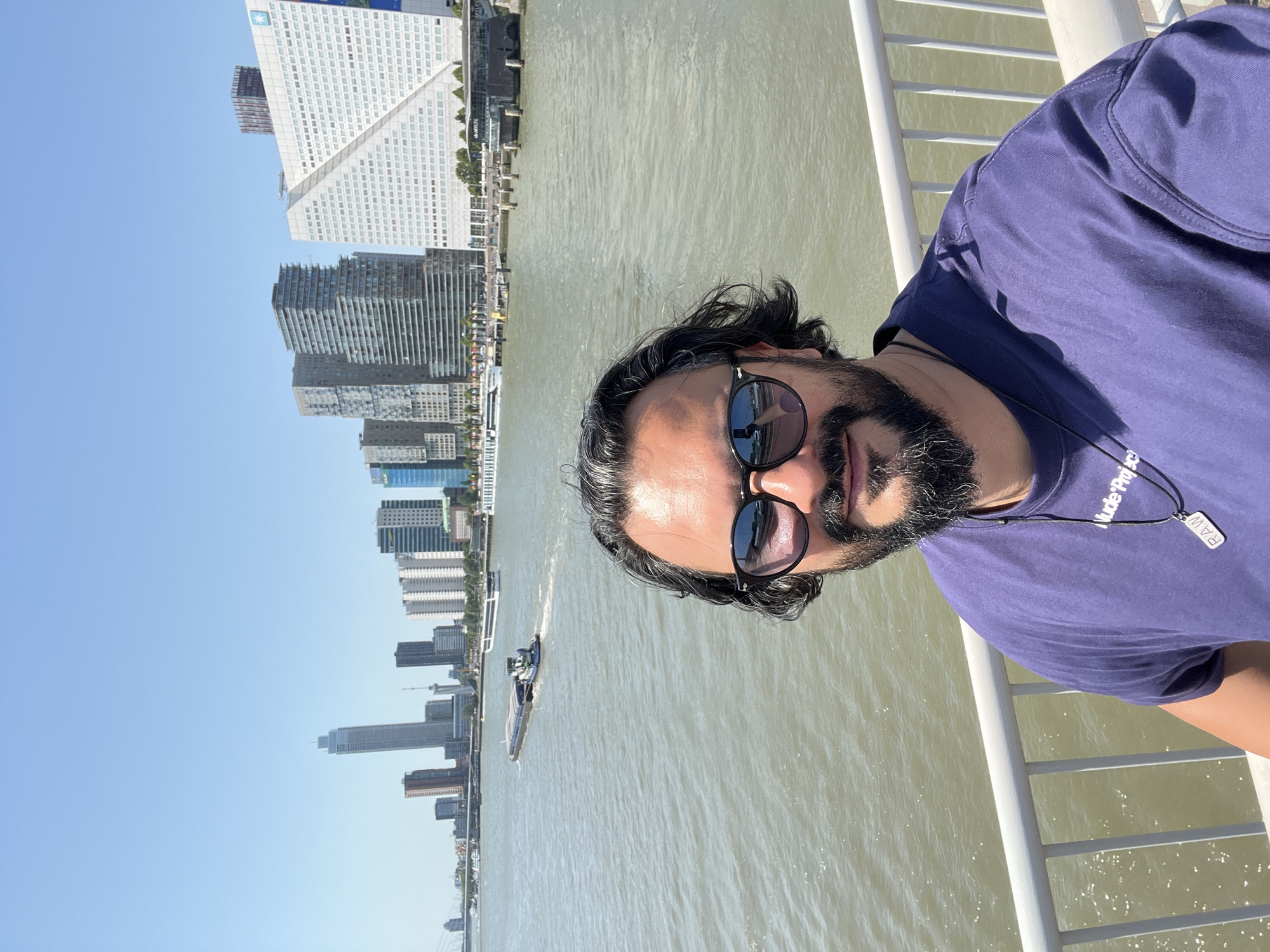 A man with dark, wavy hair and a beard, wearing sunglasses and a purple t-shirt, takes a selfie in front of the Rotterdam skyline. The skyline features modern skyscrapers and buildings, including the distinctive white building with a diagonal element. A wide river separates the viewer from the city, with a barge and a passenger boat visible on the water. The man is leaning on a white railing. The sky is clear and blue.