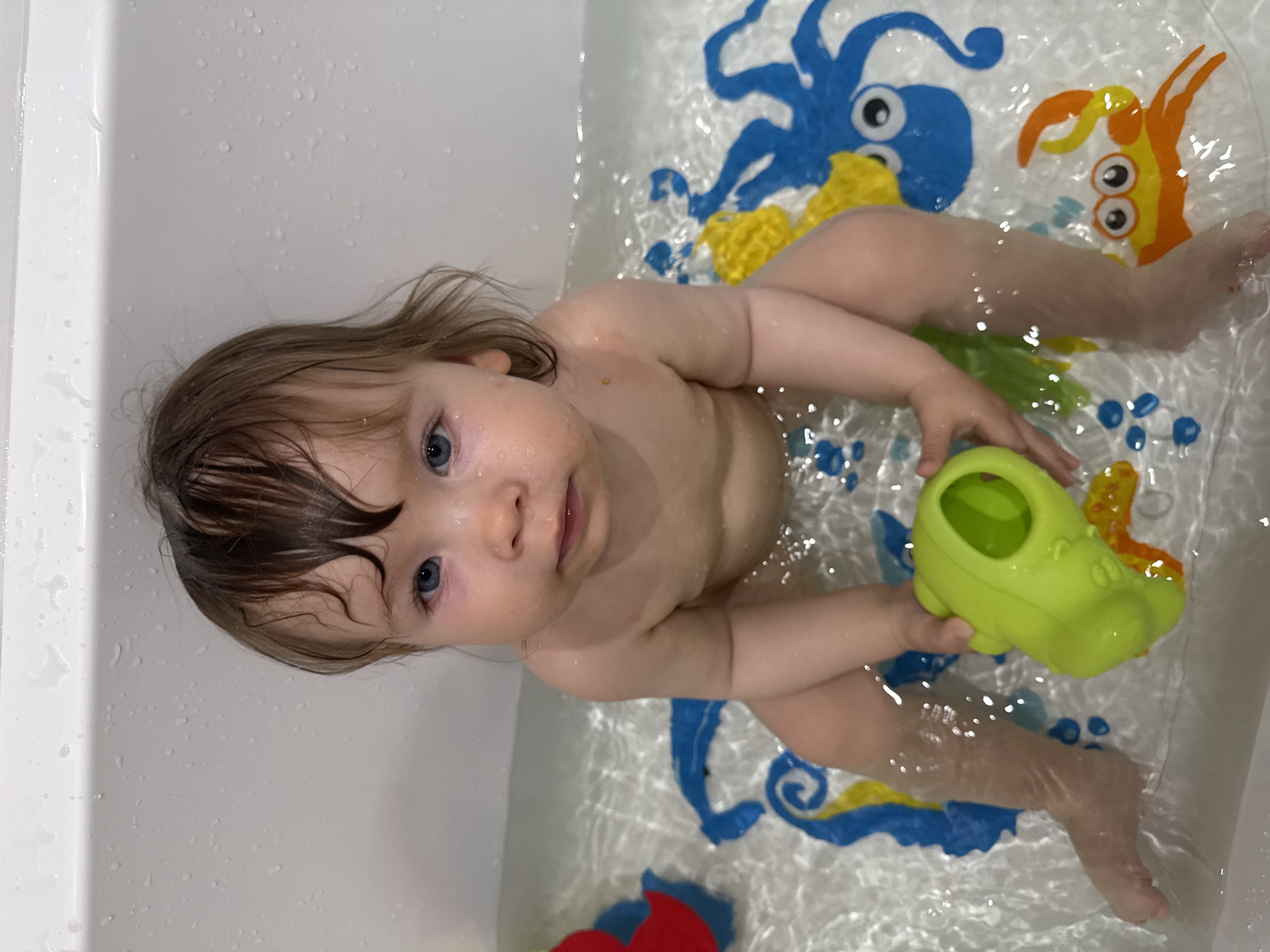 A young child is taking a bath in a bathtub. The child is lying on their back in the water, with their head tilted up and their eyes looking at the camera. The child has light brown hair and blue eyes. They are holding a green toy in their right hand.
The bathtub is white and has a non-slip mat on the bottom with colorful cartoon sea creatures, including a blue octopus and an orange crab. The water is clear and shows ripples from the child's movement. Droplets of water are visible on the side of the bathtub.
The image appears to be taken indoors during the daytime, likely in a home bathroom in Moscow, Russia. The child's expression is calm and curious. The overall scene suggests a playful and relaxed bath time. There is no visible text in the image.