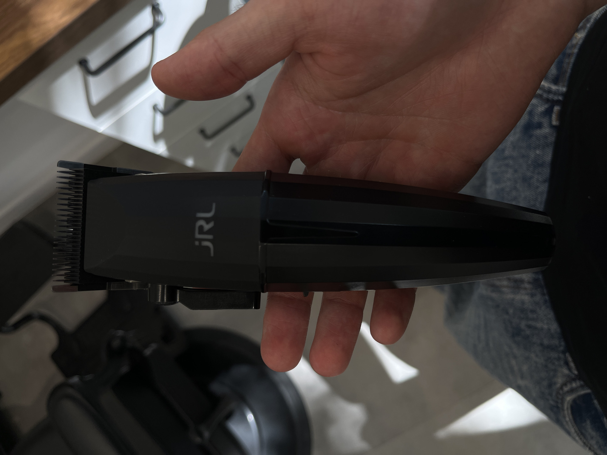 The image shows a person's hand holding a black electric hair clipper horizontally. The hand is positioned with the palm facing upwards, fingers curled around the device. The clipper is sleek and dark, with cutting teeth visible on its left side. The brand name "JRL" is prominently displayed in white text on the top surface of the clipper.
In the background, to the upper left, a light-colored, possibly white, cabinet or drawer unit is visible, featuring dark, slender, horizontal handles. A blurred, dark blue, textured fabric, resembling denim jeans, can be seen on the right side and lower right of the frame, indicating the person's attire. Below the clipper and the hand, a dark, out-of-focus object, possibly a stand or another piece of equipment, is partially visible. The scene is indoors, illuminated by artificial lighting.