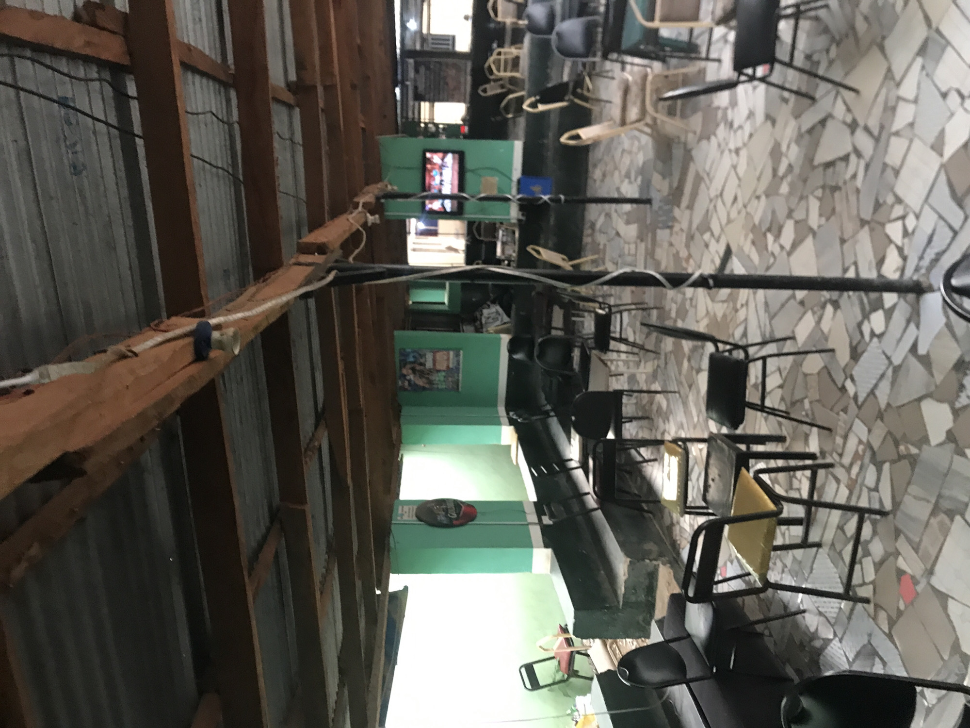 The image shows an indoor seating area that appears to be part of a restaurant or bar. The ceiling is constructed from corrugated metal and wooden beams, giving it a rustic appearance. The walls are painted a vibrant green, with white trim along the lower section. Several tables and chairs are arranged throughout the space. Some chairs are black, while others are a light beige color. A television screen is mounted on the green wall in the background, displaying an image that is too indistinct to identify. There are also posters on the walls. The floor is paved with irregularly shaped stone tiles in various shades of gray and white. In the foreground, a black pole stands vertically, possibly supporting the ceiling structure or part of a lighting fixture. The overall impression is of a casual, open-air dining or gathering space.