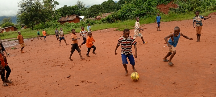 In the city of Ngalula, Democratic Republic of the Congo, a group of approximately 15 children is engaged in a game of soccer on a reddish-brown dirt field. The field is expansive, showing an uneven surface with scattered debris and a small muddy area visible on the far left. The children, appearing to be male and ranging from roughly 6 to 12 years old, are dispersed across the playing area.
One child, centrally positioned in the mid-foreground, wears a red, white, and blue striped short-sleeved shirt and blue shorts, and is barefoot while actively dribbling a yellow and green striped soccer ball with his right foot. To his right, another child in a blue tank top and tan shorts, wearing dark sandals, is running forward with a bent posture. Other children are scattered throughout the scene; some are running, others are standing and observing. Most children are barefoot, though a few wear sandals or light athletic shoes. Their attire consists of t-shirts, tank tops, and shorts in various colors including orange, blue, and patterned designs.
In the background, behind a narrow strip of green grass, several small, rustic structures with reddish-brown walls and dark roofs are visible. These buildings appear to be simple, single-story dwellings. Beyond these structures, a dense expanse of dark green tropical vegetation covers a rising landform, possibly a hill or mountain, under an overcast sky.
