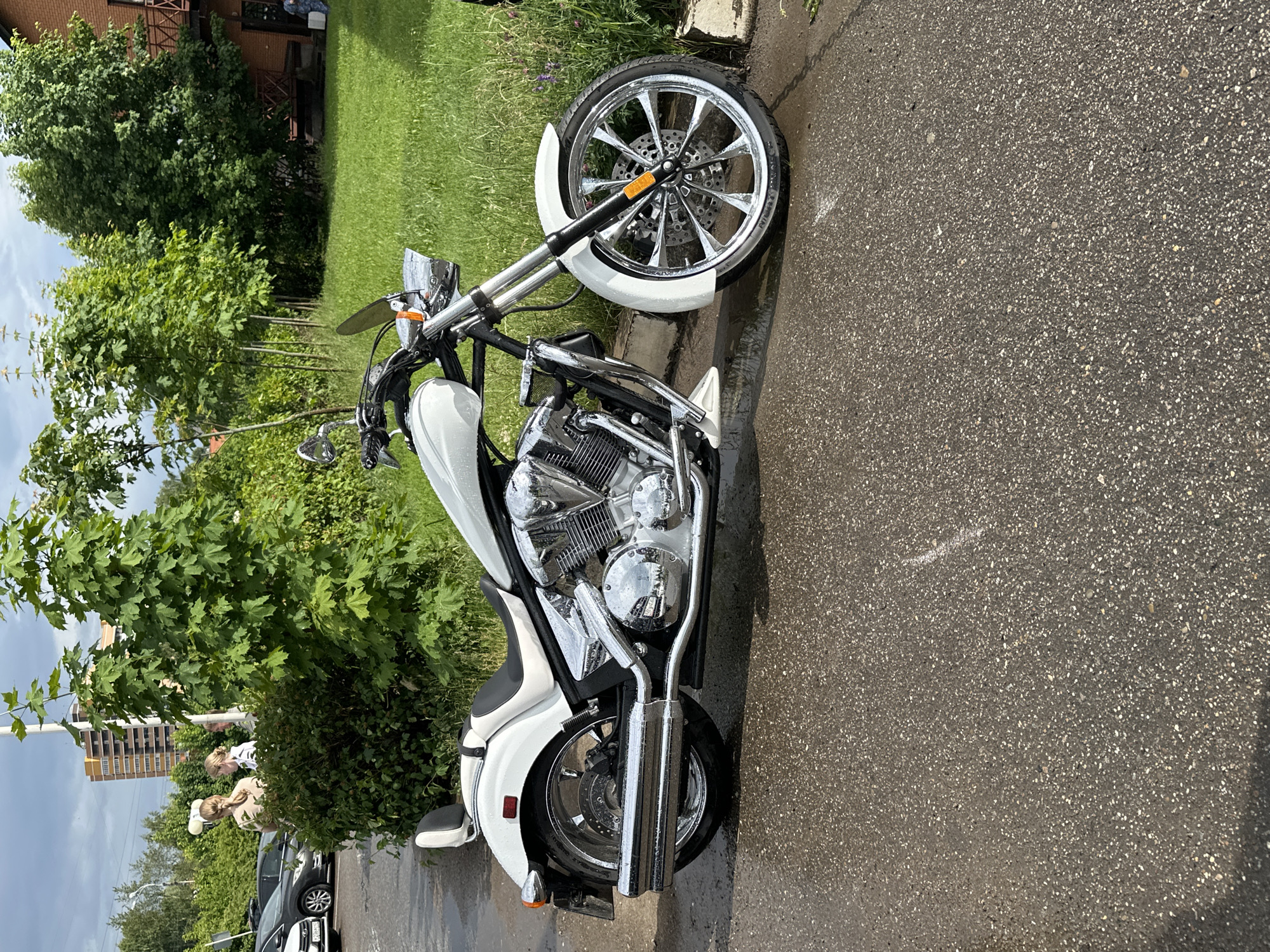 A white motorcycle is parked on a wet asphalt surface. The motorcycle has a chrome engine and exhaust pipes, and a black seat with a white passenger backrest. In the background, there is a green lawn, trees, a building with a red brick facade, and a tall residential building. Several cars are parked in the distance. The sky is overcast. There are three people standing near some bushes to the left of the motorcycle. The image appears to have been taken on a cloudy day in Izhevsk, Russia.