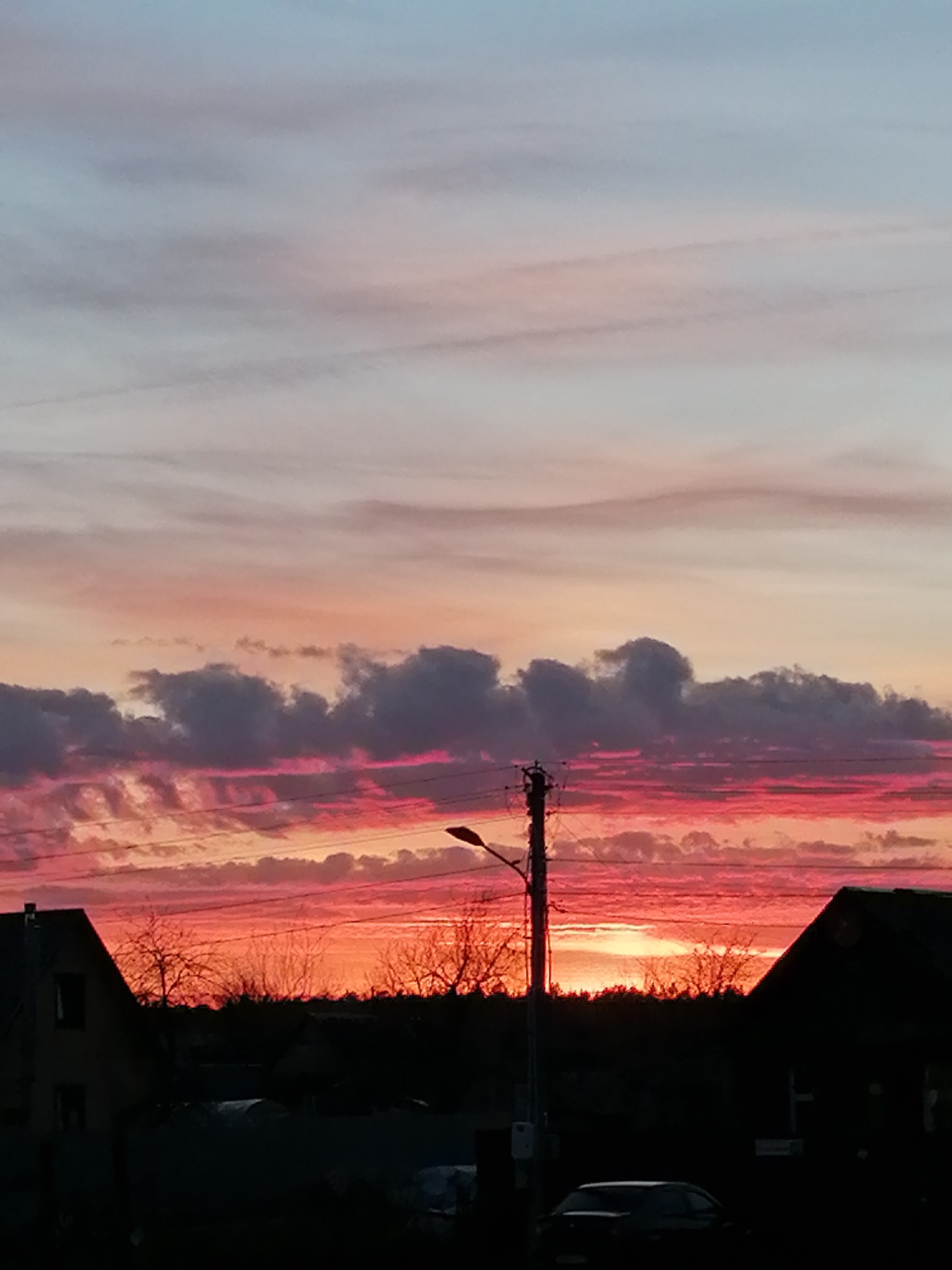 The image depicts a vivid sunset in a residential area. The sky exhibits a gradation of colors, from pale blue at the top to intense shades of pink and red near the horizon. A band of dark, puffy clouds lies in the middle ground, reflecting the sunset's colors. Silhouetted against the colorful sky are the rooftops of houses on either side, and bare trees and a power pole with a streetlight in the middle ground. A car is visible in the foreground, parked on what appears to be a street. The location appears to be a suburban neighborhood.