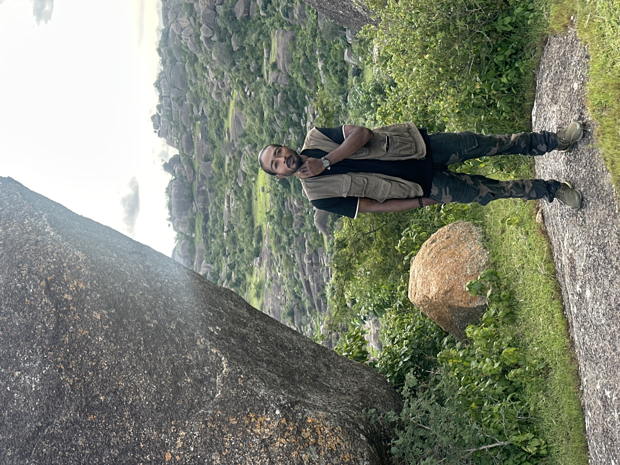 A man stands on a rocky outcrop in a semi-arid, hilly landscape near Jos, Nigeria. He is positioned slightly left of center, facing the camera. He wears a black t-shirt, a tan utility vest with multiple pockets, camouflage pants, and green athletic shoes. His left hand is raised to his chin, and he wears a watch on his left wrist.
The foreground is dominated by a large, rough-textured grey boulder to the left, obscuring part of the background. In front of the man, a smaller, reddish-brown boulder sits amongst lush green vegetation and grass. The middle ground and background are characterized by rolling hills covered in more grey boulders of various sizes and scattered green shrubs and trees. The sky is overcast with some clouds visible. The overall impression is of a rugged, natural environment.
