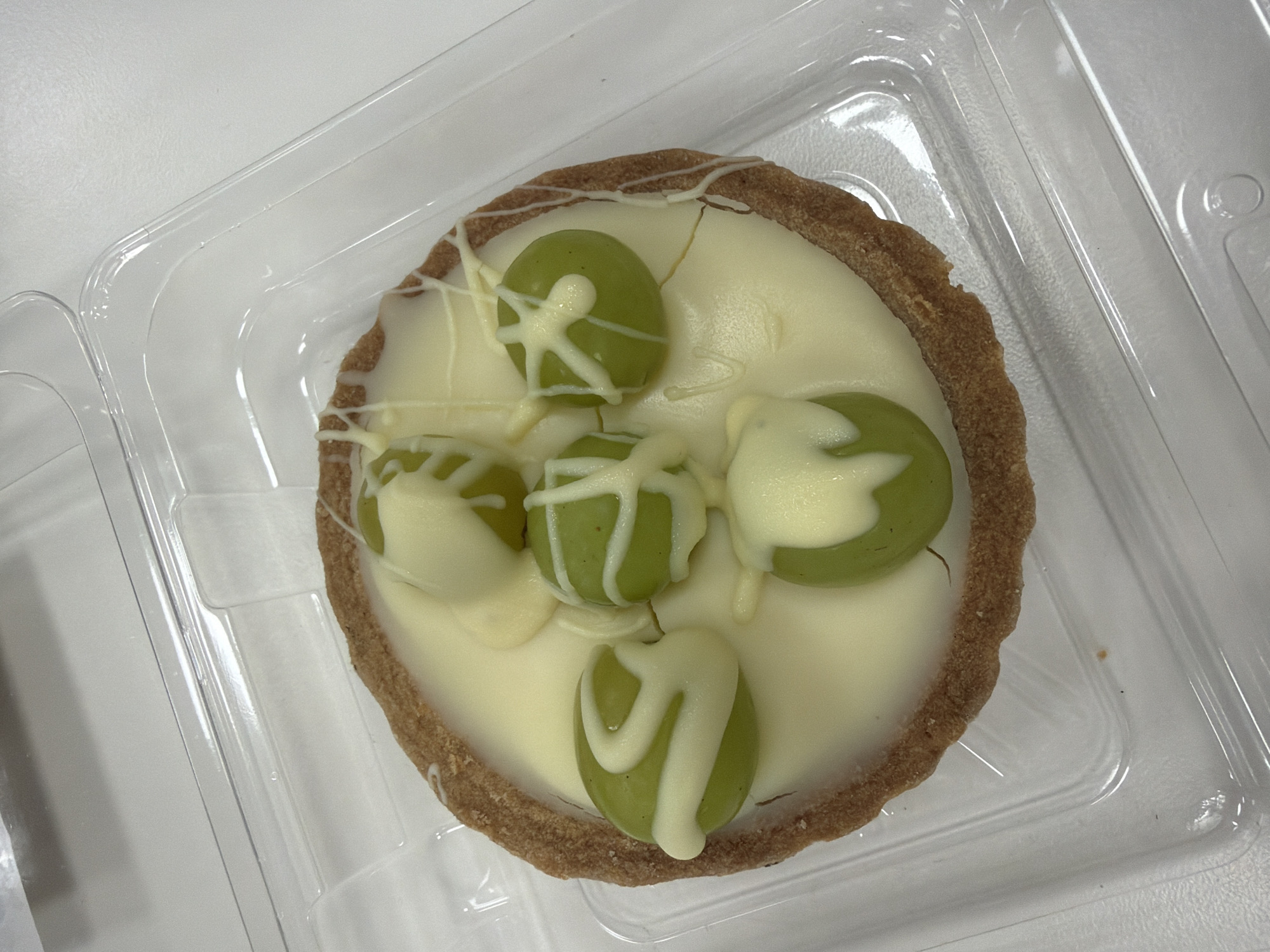 A tart with a brown crust, white filling, and green grapes drizzled with white chocolate is positioned inside a clear plastic container. The container rests on a white surface. The location is unknown.