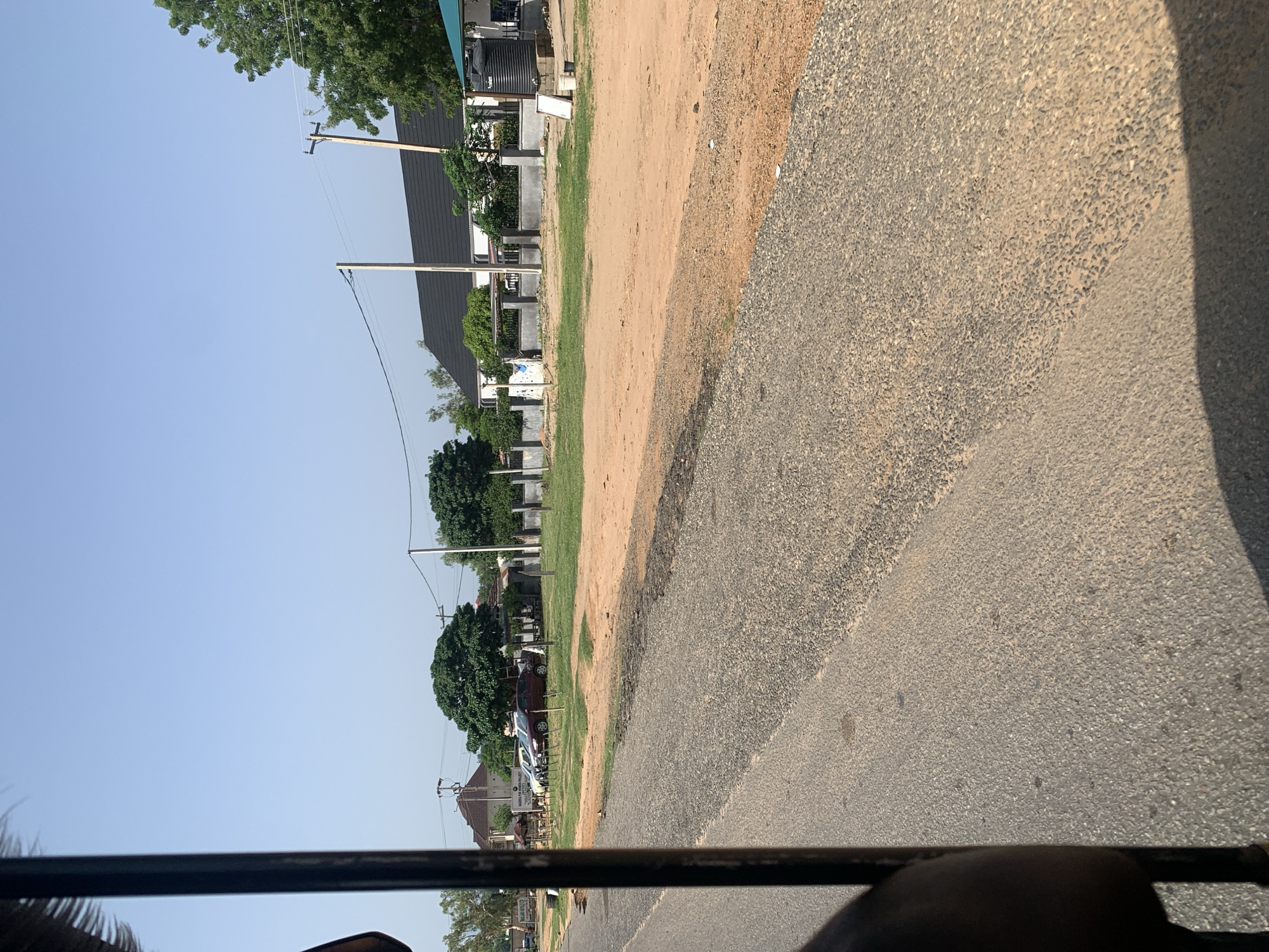 An outdoor, eye-level shot captures a scene along a paved road, framed by a dark, vertical bar on the left, suggestive of being taken from a vehicle. The road's surface is a mix of grey asphalt and lighter-colored gravel, with a sandy dirt patch bordering it on the right. Beyond the dirt, a grassy area hosts several parked cars, including a white sedan, a silver car, and a red sedan. A large, mature tree with a full canopy stands prominently behind the cars. Further back, a white building with a dark, pitched roof is visible, along with more greenery and telephone poles supporting power lines. The sky is a clear, pale blue, indicating a sunny day. The scene is devoid of people.