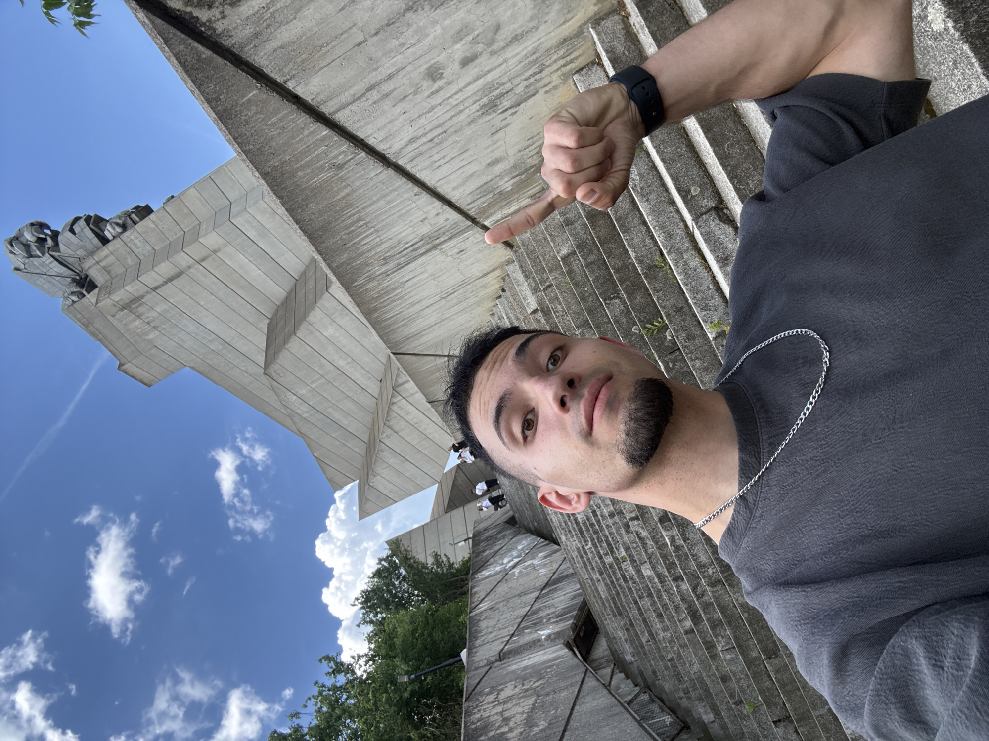 A low-angle selfie taken outdoors in Shumen, Bulgaria, showcases a man with dark hair, fair skin, and a trimmed beard, wearing a grey t-shirt and a silver chain necklace, pointing his index finger towards the Monument to 1300 Years of Bulgaria. The monument is constructed of light grey concrete, with a sculptural element at the top against a blue sky with scattered clouds. In the background, trees and vegetation are visible, and a set of concrete stairs leads to the monument.
