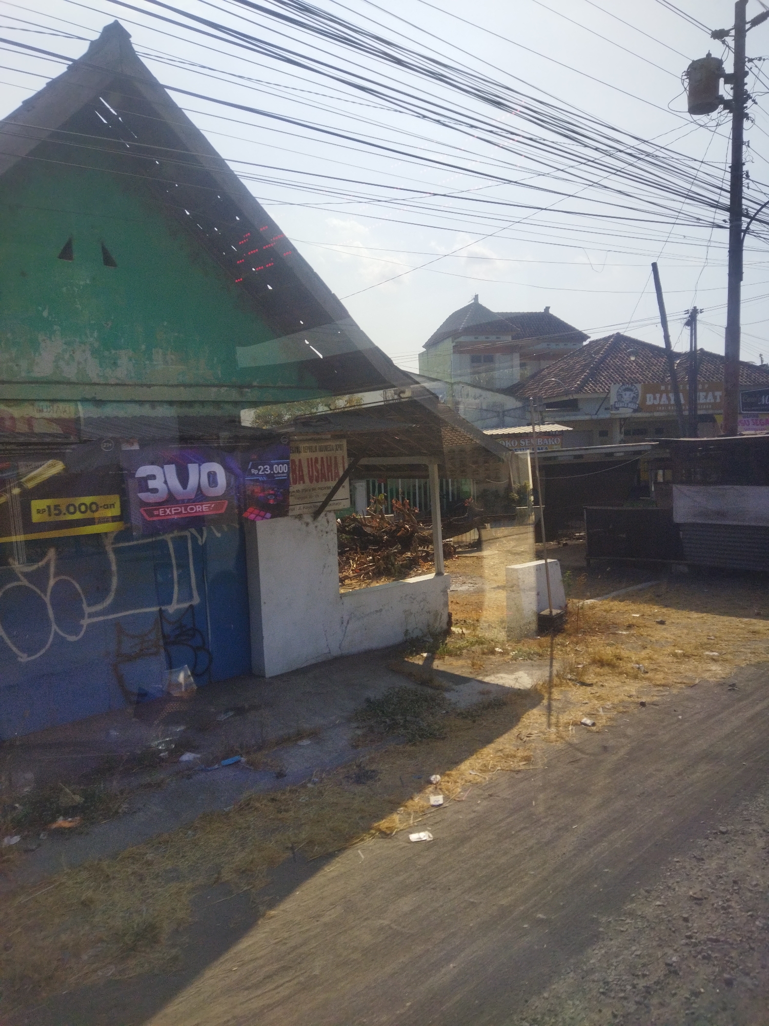 The image shows a street scene in Nguter, Indonesia. A green, dilapidated building with a triangular roof is visible on the left, with red lights forming a pattern on its side. A blue wall with graffiti is in front of the building. A banner with the text "3VO EXPLORE?" and "RP 15.000-on" is displayed. Another banner reads "BA USAHA" and "REPUBLIK INDONESIA". Several power lines crisscross the sky above the scene. In the background, buildings with tiled roofs are visible, along with a utility pole with a transformer. The ground is covered with dry grass, dirt, and some litter. The lighting suggests it is daytime.