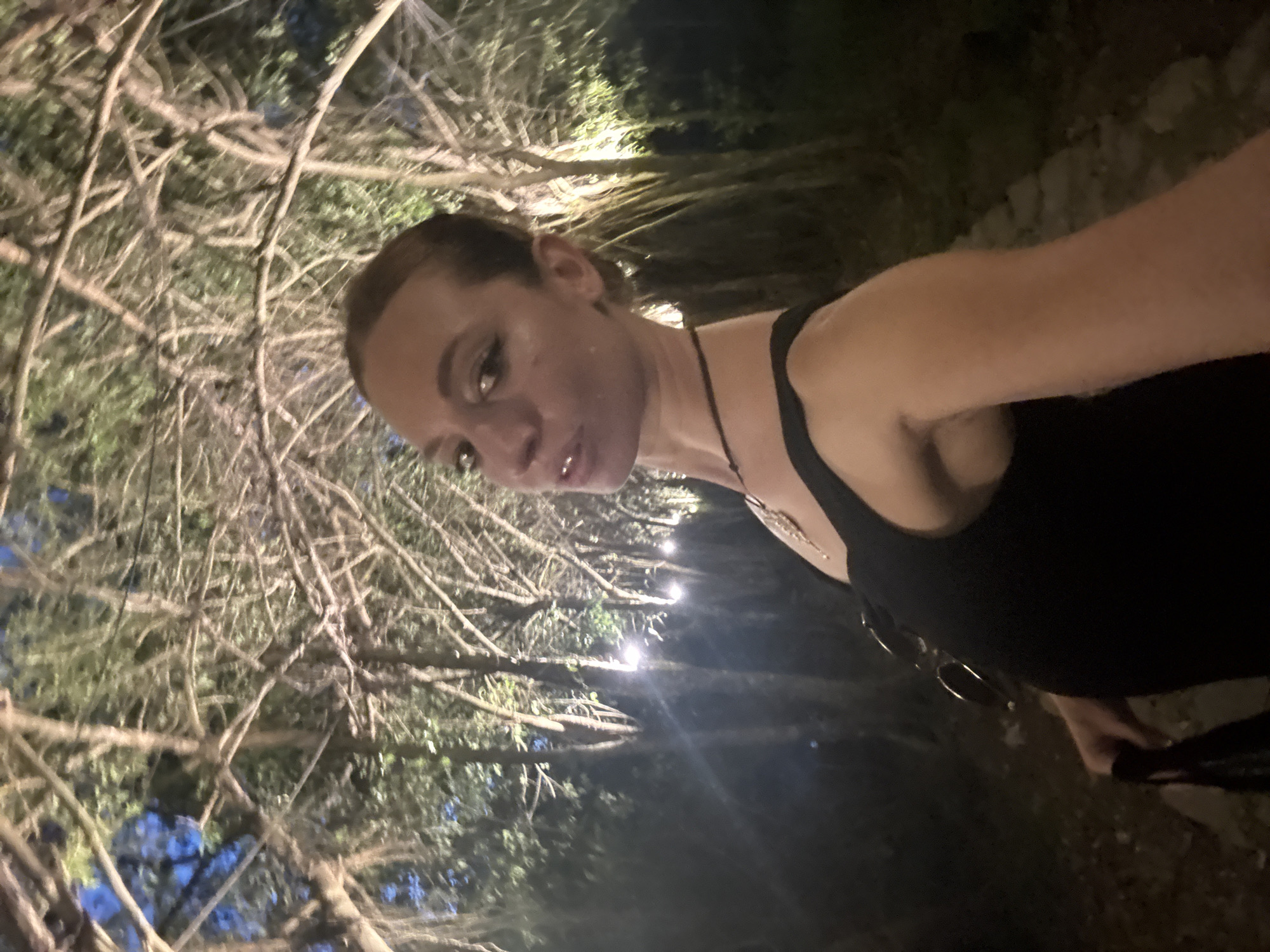 The image shows a young woman in a black sleeveless top, taking a selfie at night. She has dark hair pulled back and is wearing subtle makeup, including winged eyeliner. She is wearing a necklace with a pendant and has a septum piercing. The background is a dimly lit forest or park area, with trees and branches arching overhead. Lights are visible among the trees, suggesting the area is illuminated for evening access. The ground appears to be a path made of stones or gravel. The woman is holding a dark bag in her left hand. The lighting creates a somewhat ethereal and mysterious atmosphere. The specific location within Nam Hoa Lư Ward, Vietnam, is not identifiable from the visual information in the image itself.